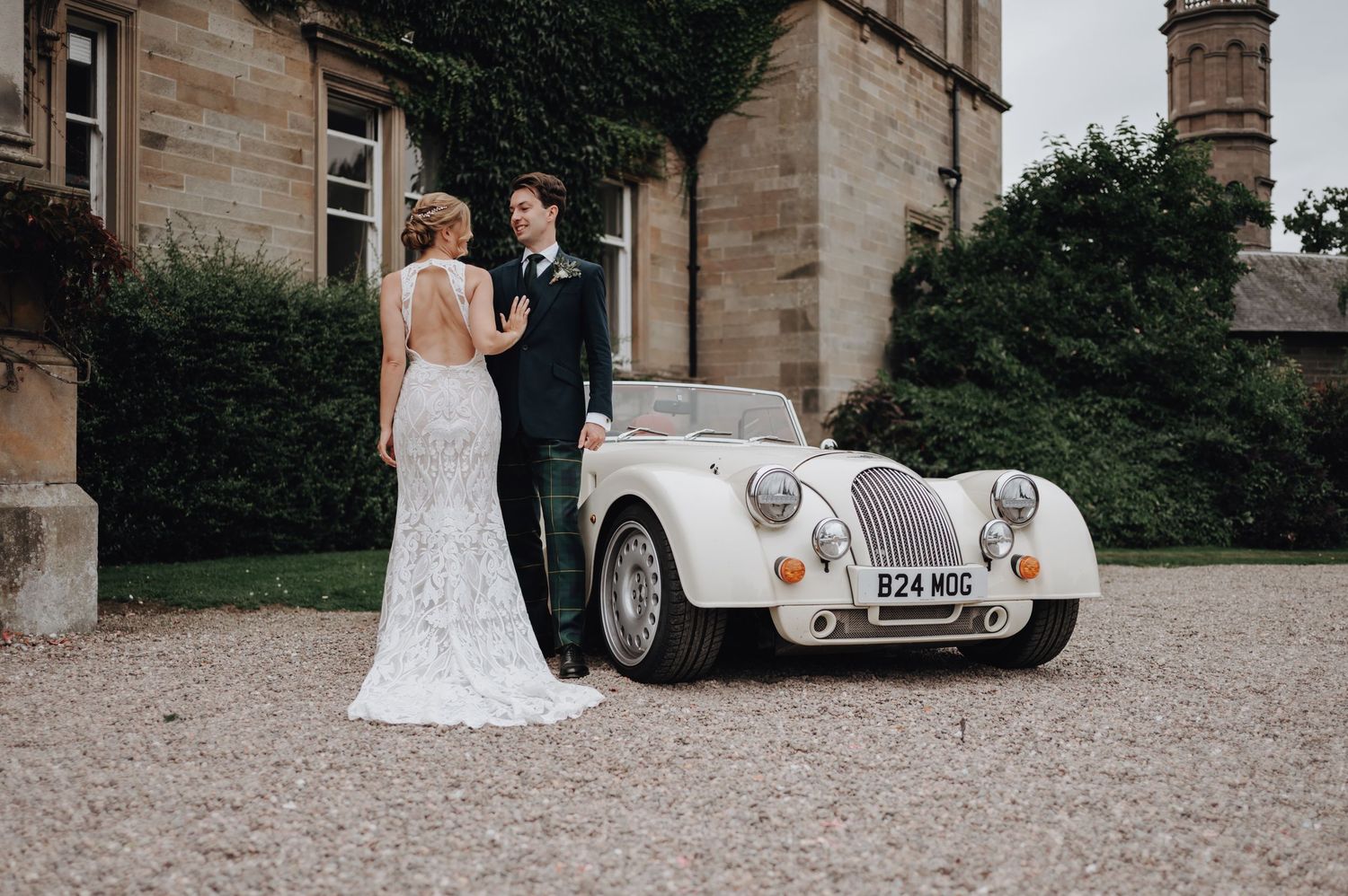 Classic vintage wedding car parked outside stone building with couple in formal wedding attire beside it.