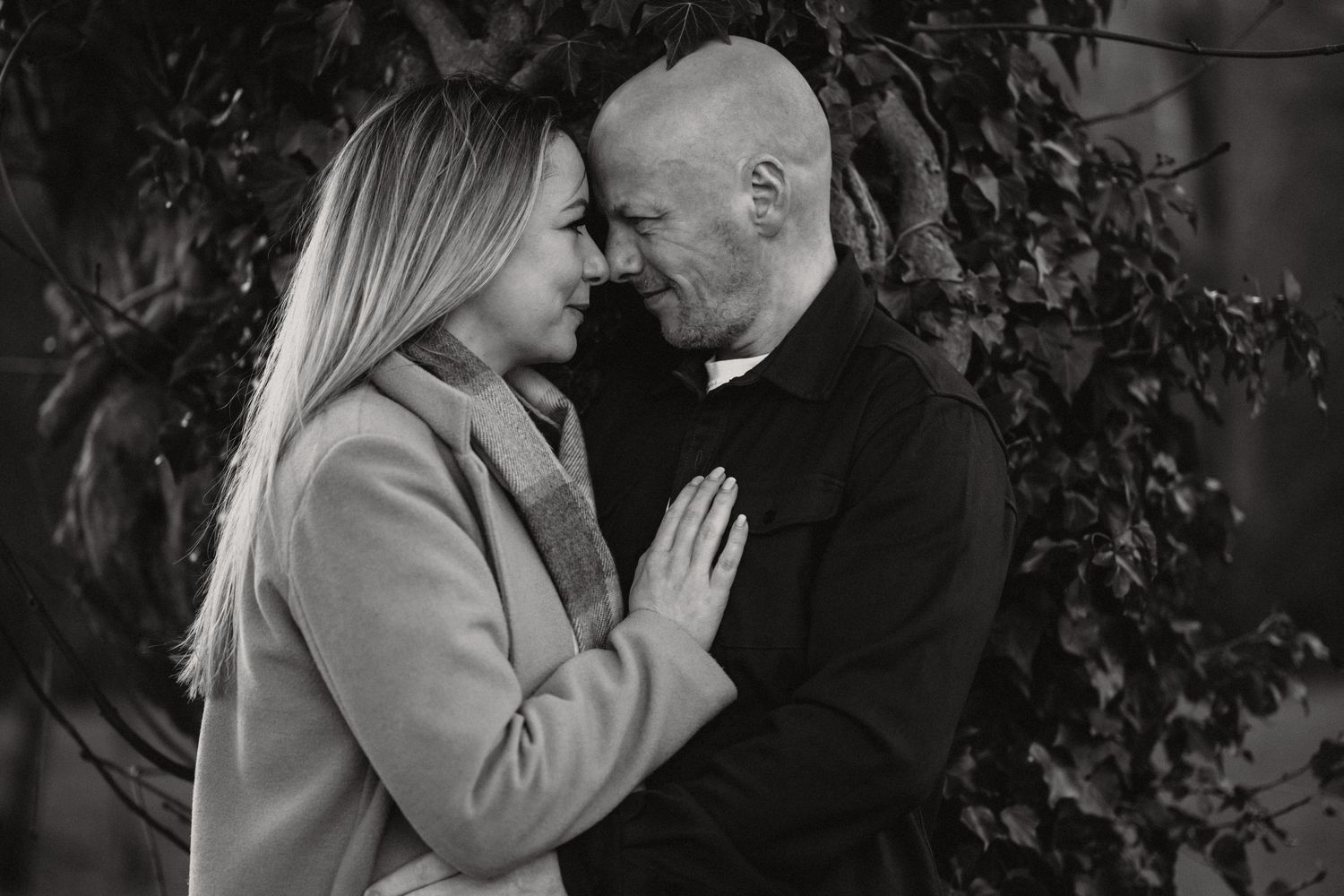 Black and white portrait of couple in tender embrace against leafy background.
