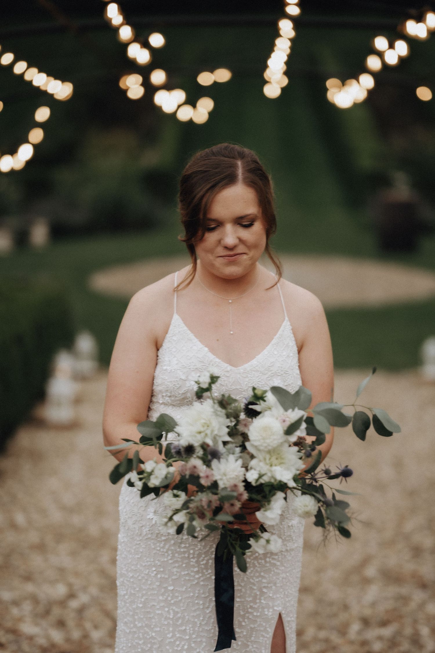 Bride holds elegant white and green bouquet while standing under string lights.
