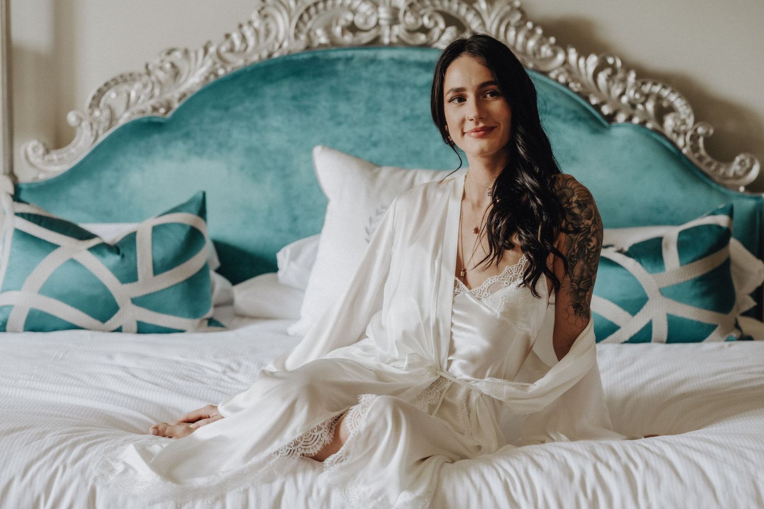 Woman in white robe sitting on bed with turquoise tufted headboard during bridal preparation.