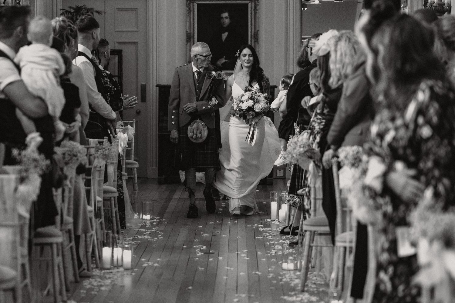 A wedding ceremony aisle lined with candles as the bride enters with her escort in a traditional Scottish venue.