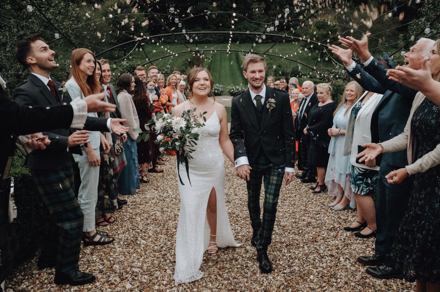Happy couple exits ceremony while guests throw confetti under string lights.