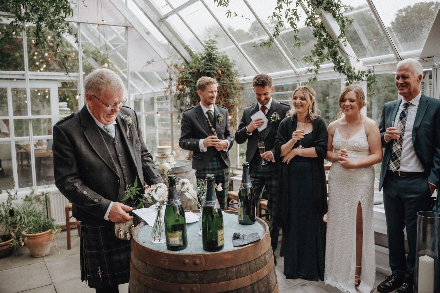Wedding guests gather around a wine barrel in a greenhouse setting while enjoying drinks and conversation.