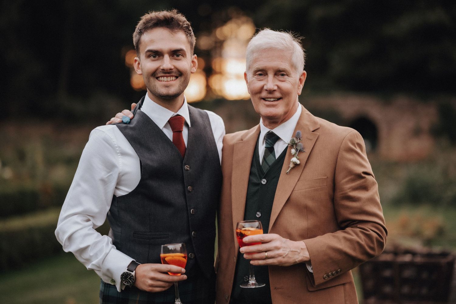 Two guests enjoying drinks at a formal event with one wearing a tan suit and the other in a vest and tie.