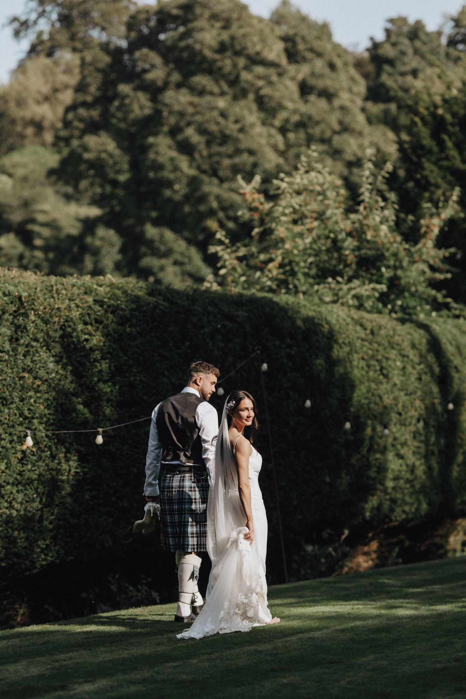 Newlyweds walking together on a manicured lawn with hedges in the background.