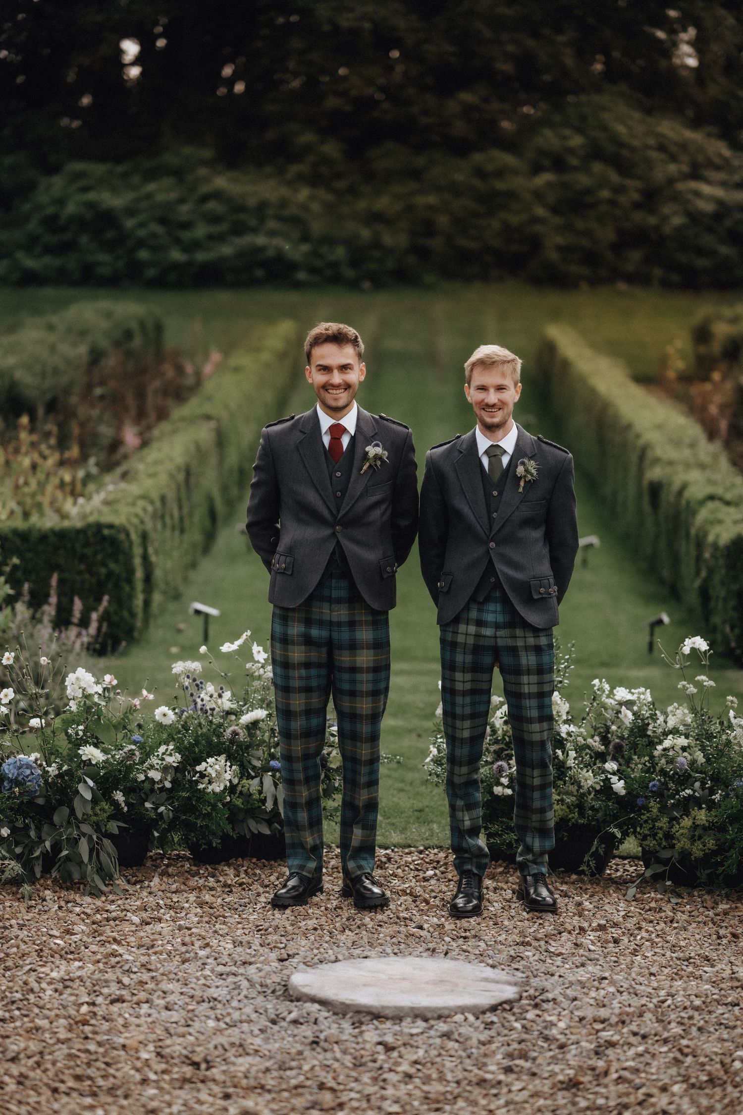 Two people in matching formal wear with tartan kilts standing in a manicured garden.