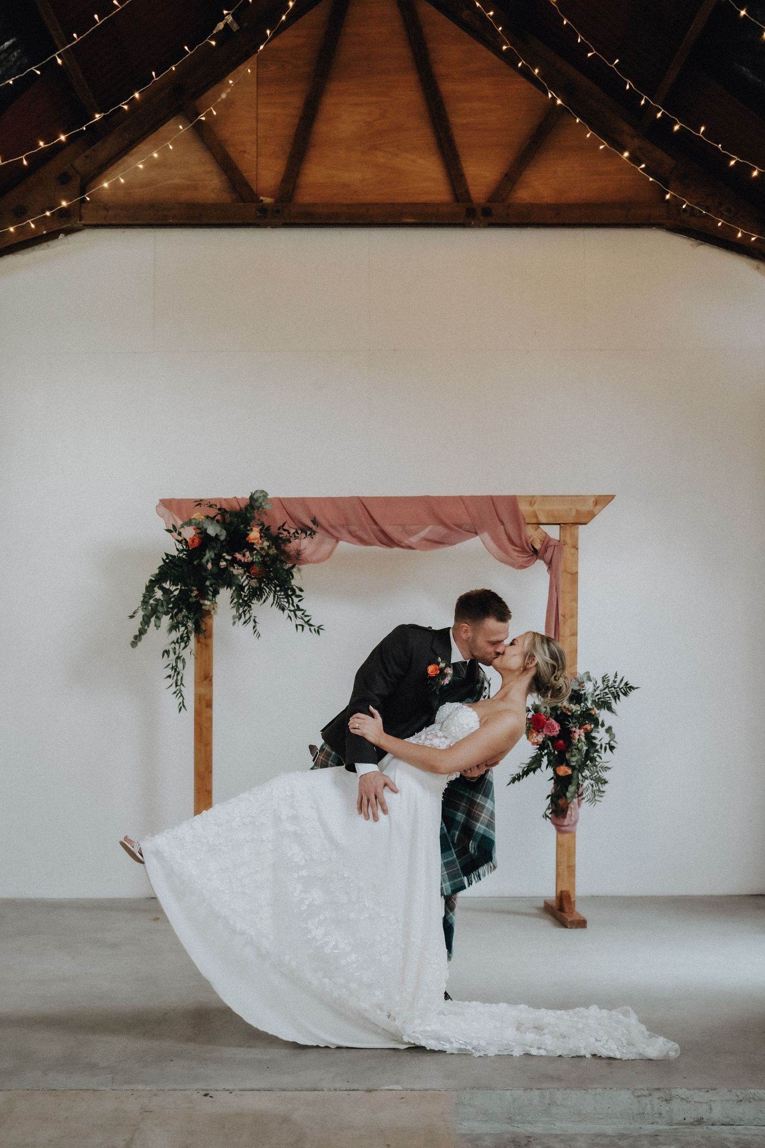 Romantic dip kiss under wooden arch decorated with pink fabric and greenery.