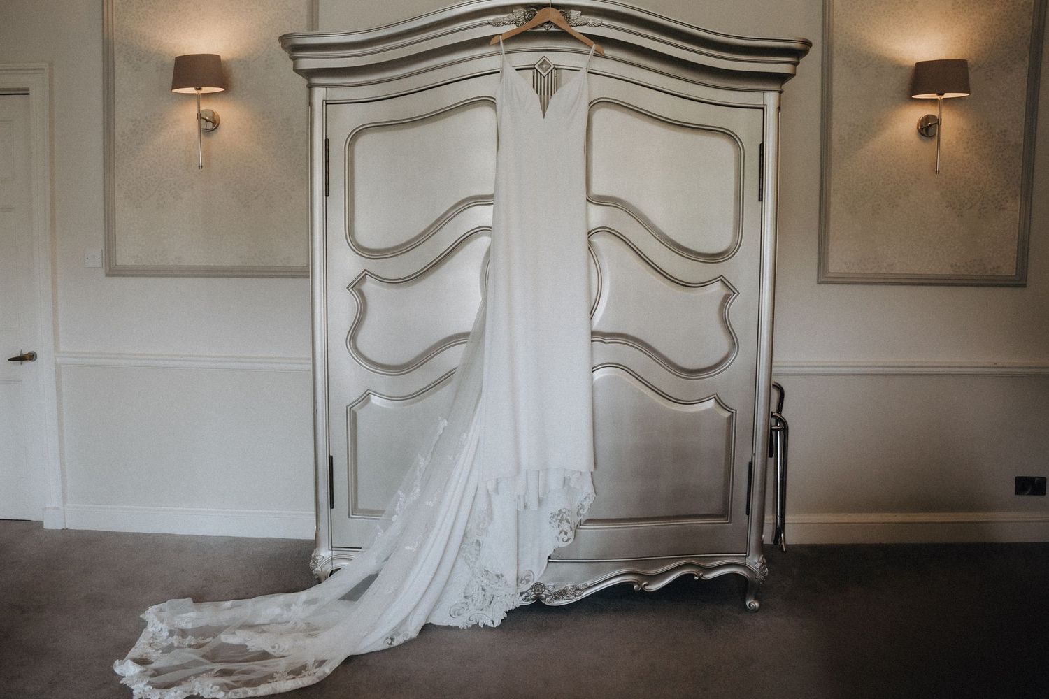 An elegant white wedding dress displayed in front of an ornate white vintage wardrobe.