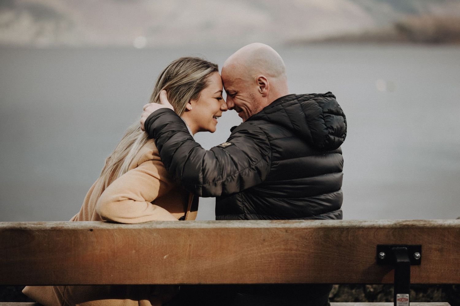Romantic couple embracing on a bench against a moody mountain landscape backdrop.