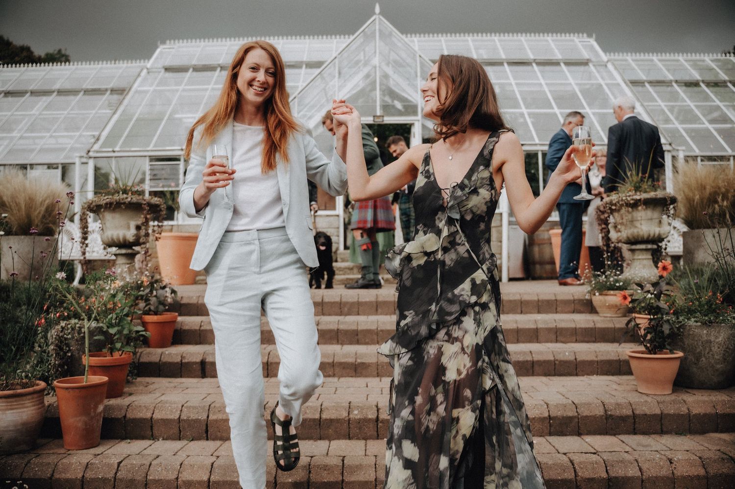 People celebrate near a Victorian-style greenhouse with potted plants on brick steps.