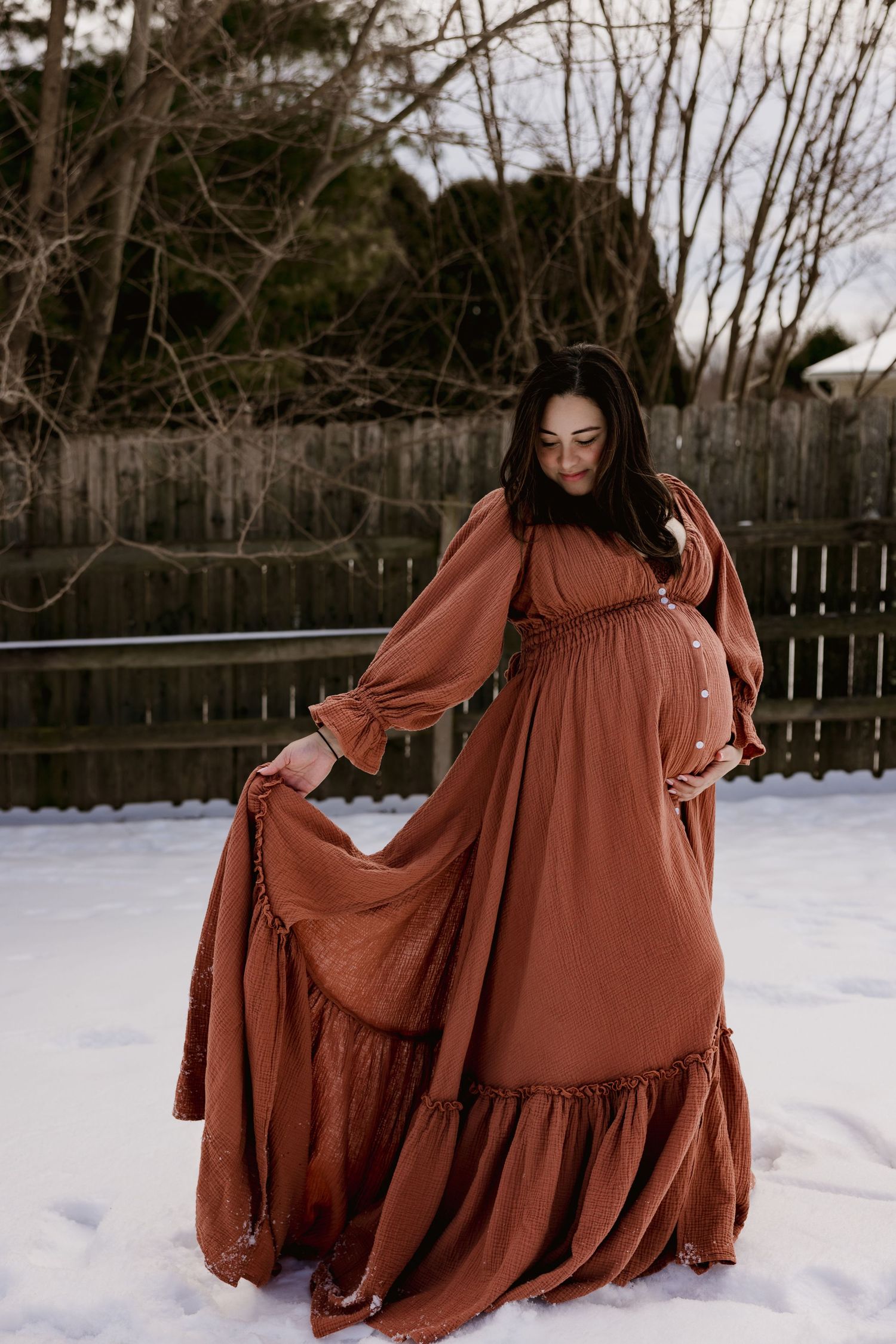 Pregnant woman in a flowing rust-colored maxi dress poses gracefully in a snowy backyard.
storytelling photographer