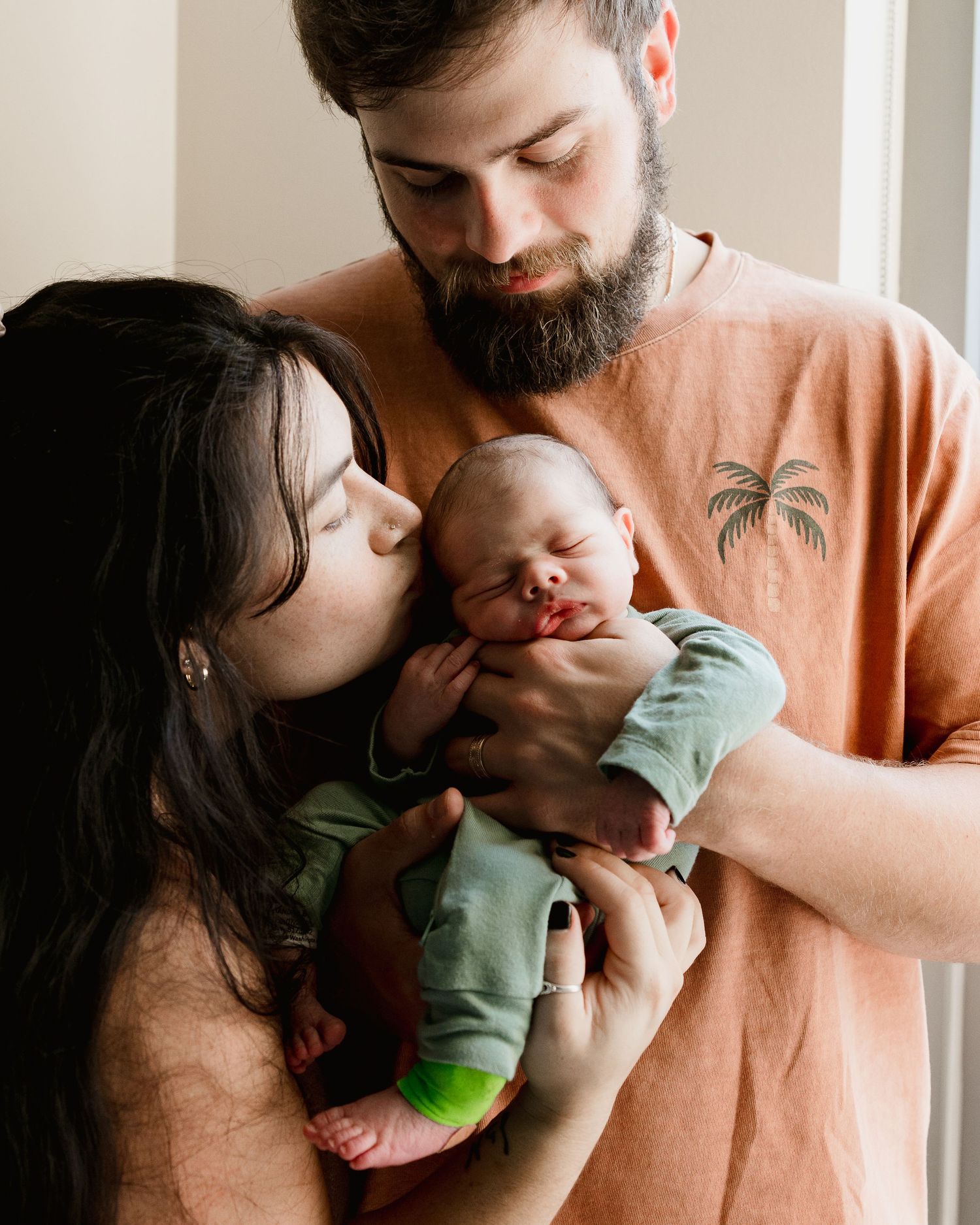 A bearded father holds a sleeping newborn while the dark-haired mother gently kisses the baby's head.