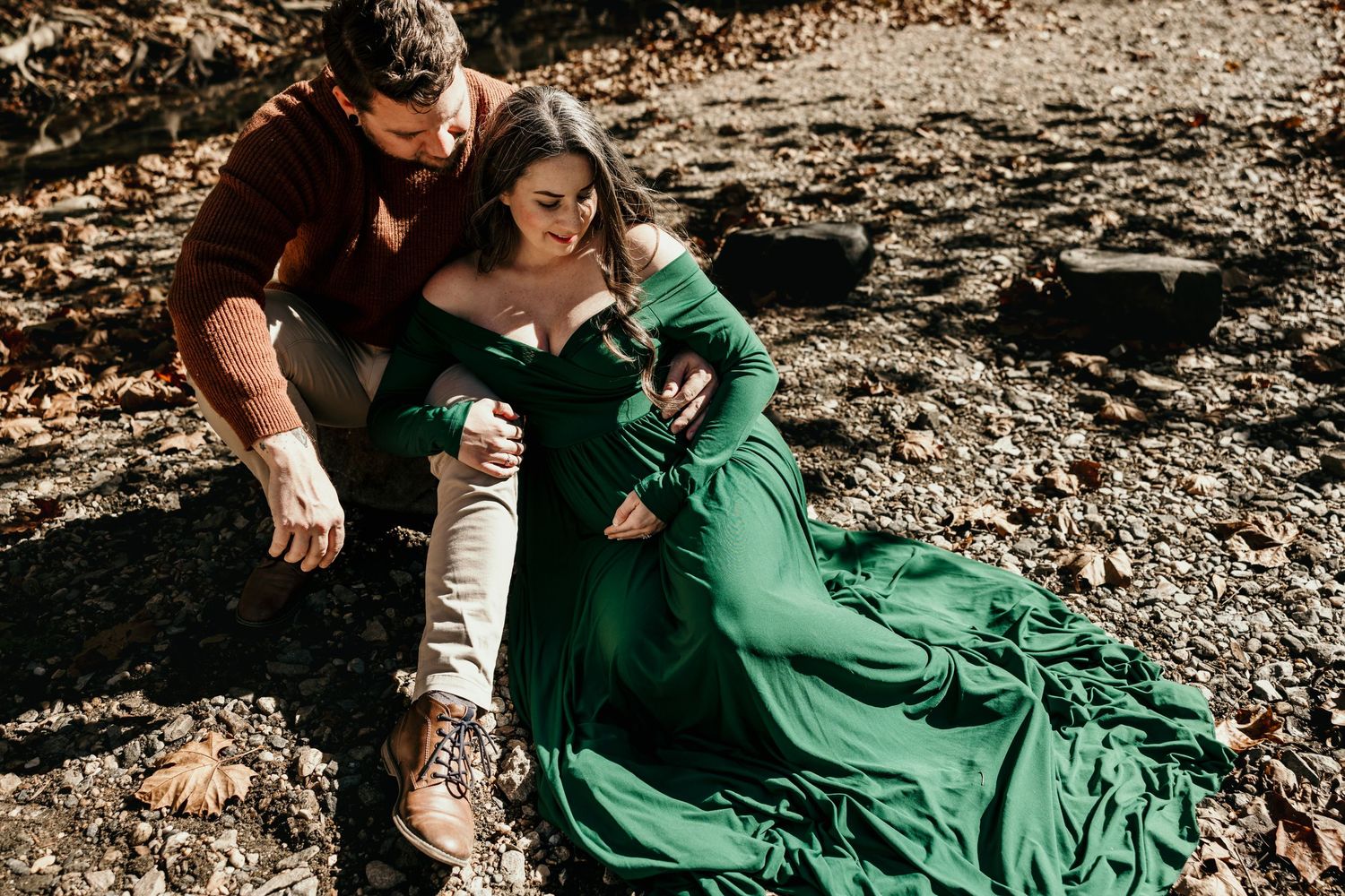 Couple sharing tender moment sitting on rocky ground, pregnant woman in emerald green gown with flowing train.