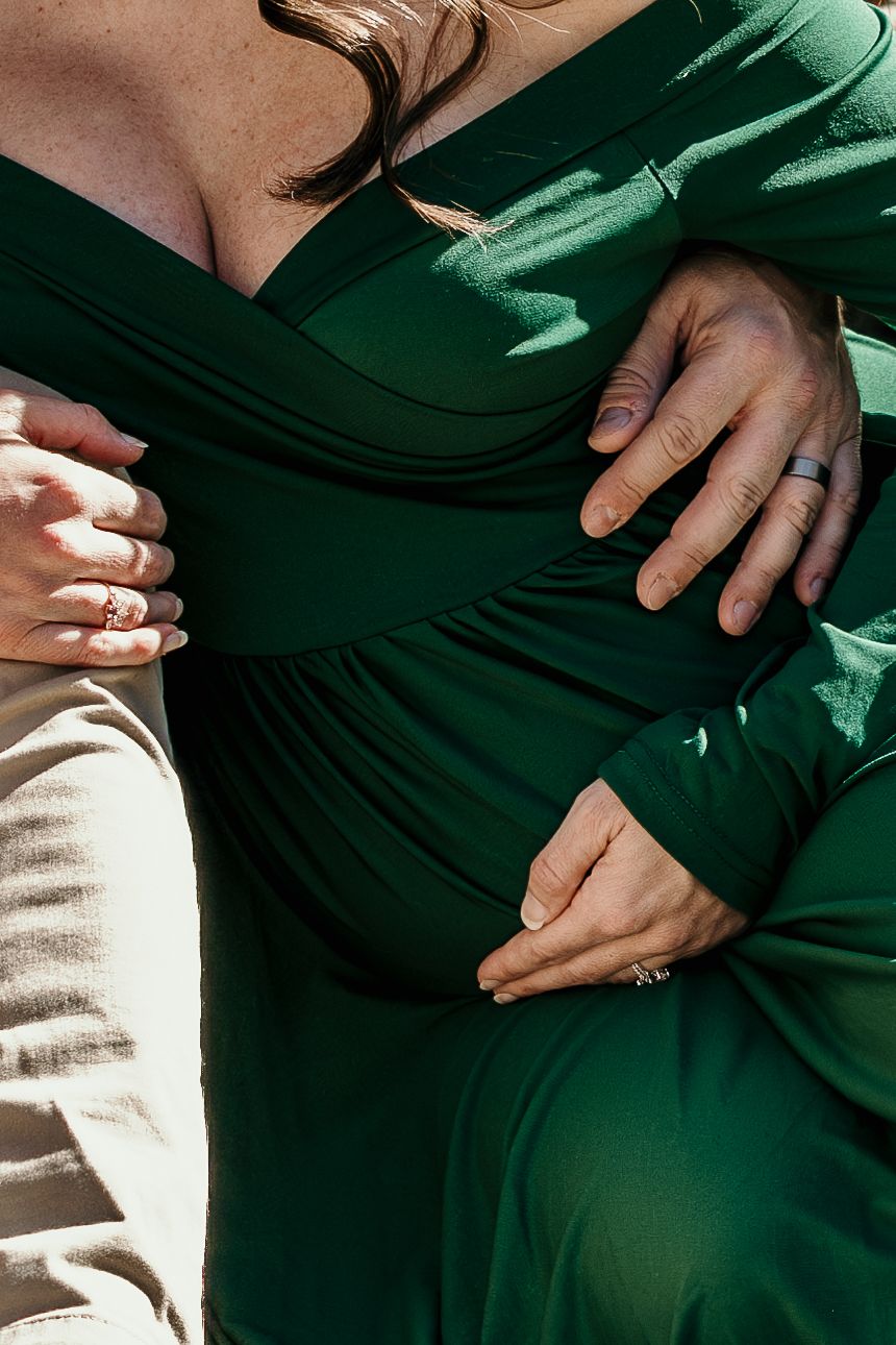 Close-up of couple cradling pregnant belly, woman in emerald green maternity dress, rings visible on hands.