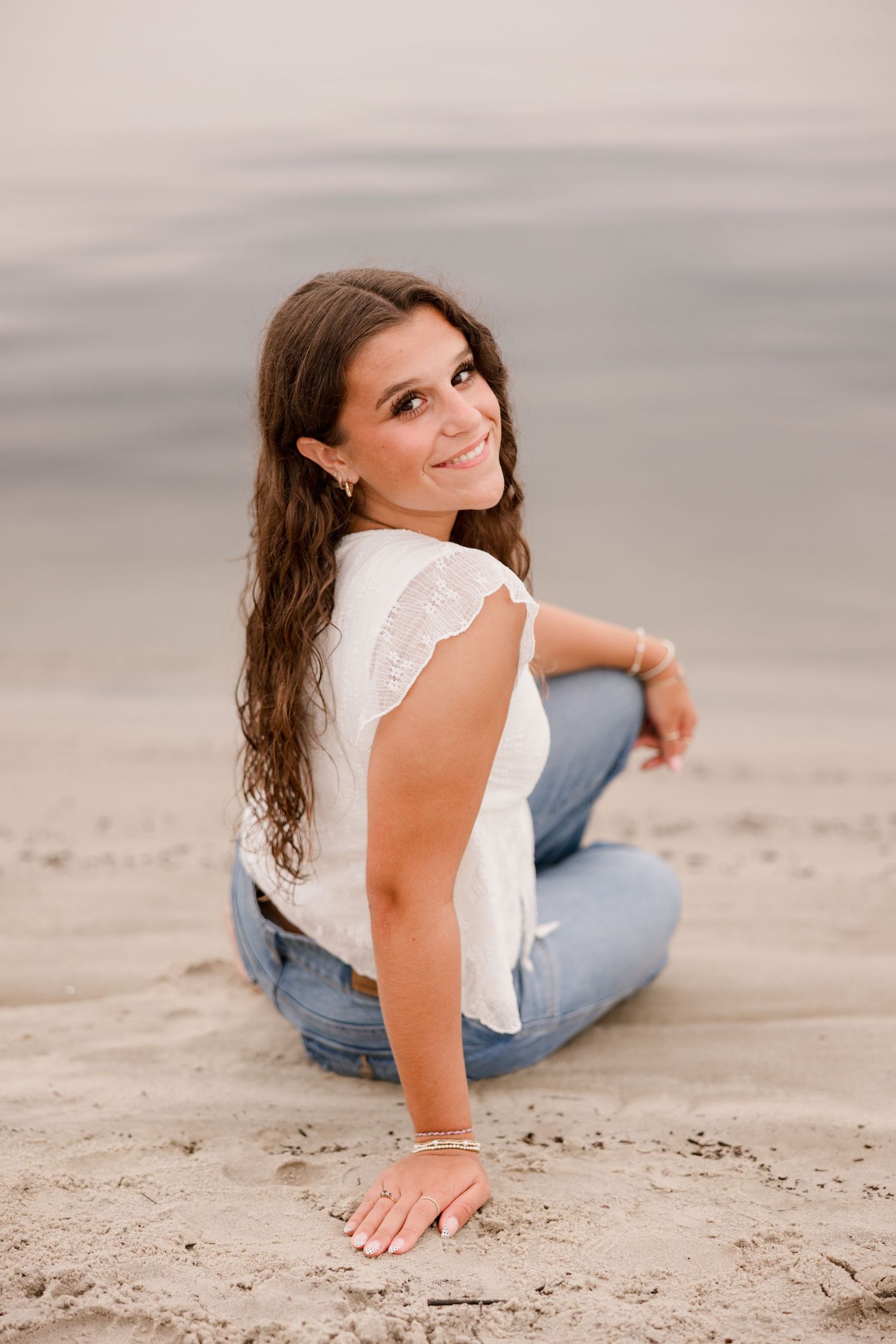 Young woman with long brown hair sits cross-legged on sandy beach, smiling over shoulder, wearing white top and jeans.