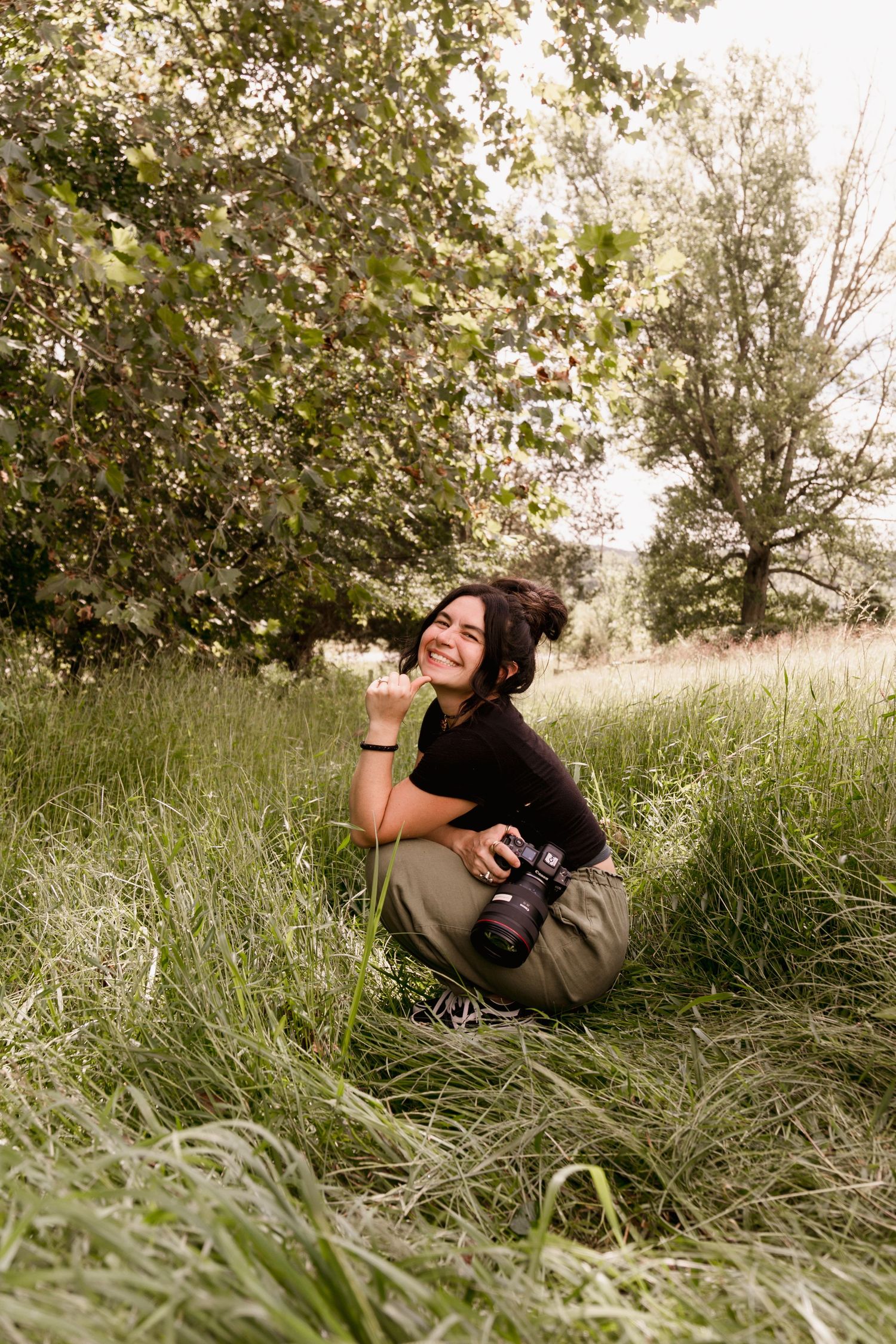 A smiling woman crouches in tall green grass holding a camera, surrounded by lush trees in a natural outdoor setting.