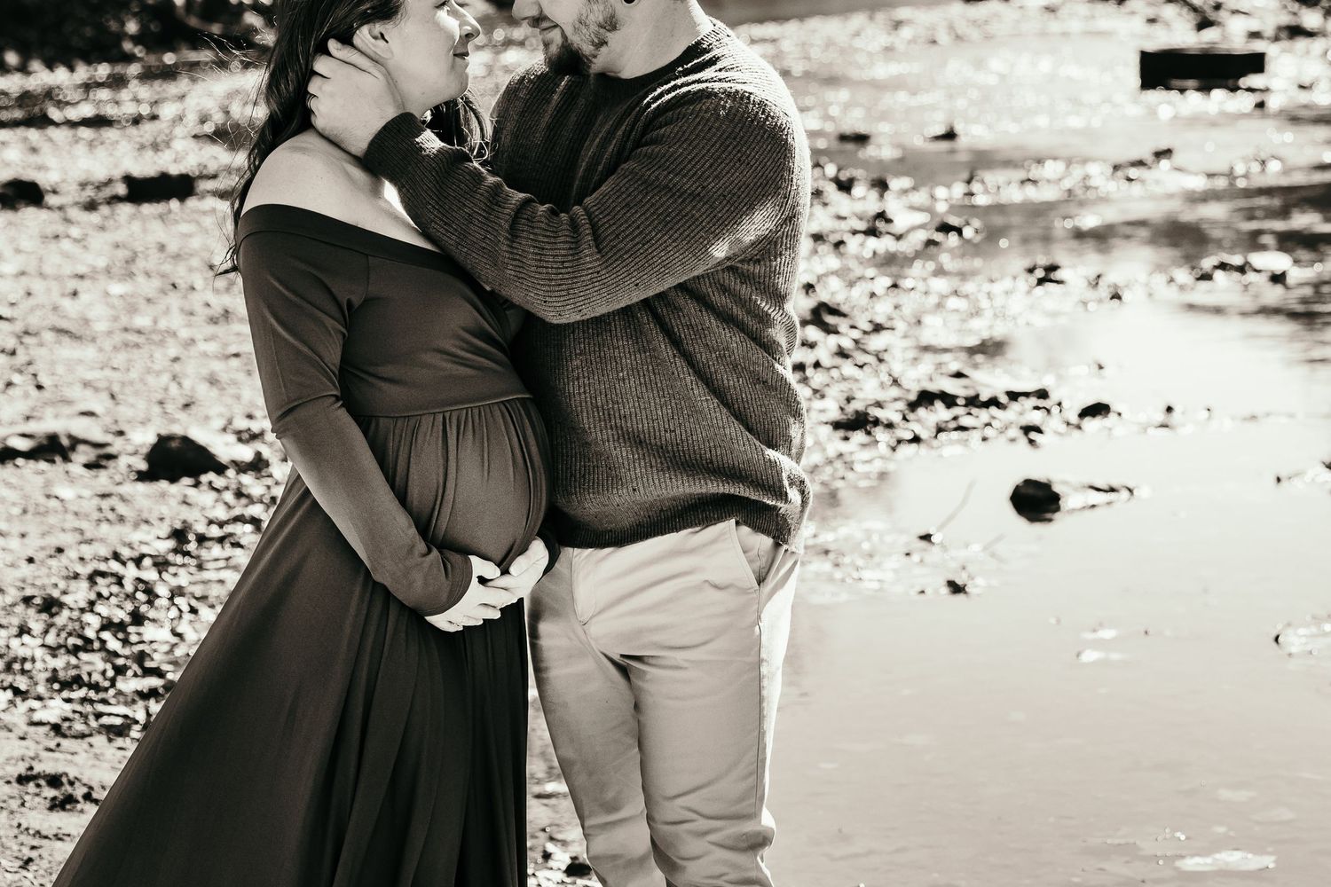 Couple embracing tenderly outdoors near rocky creek, pregnant woman in dark gown, black and white photo.