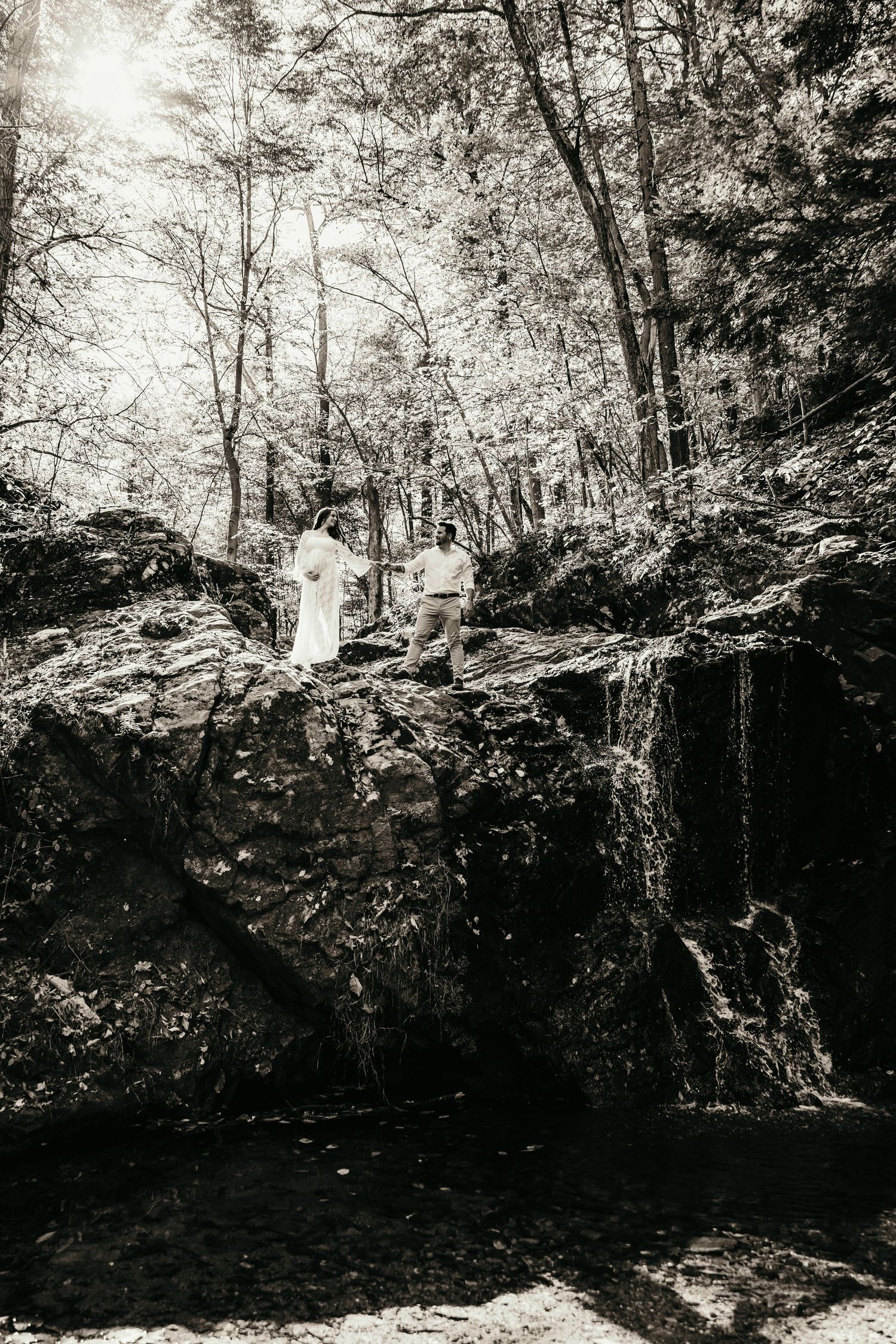 Black and white photo of couple standing on rocks near a waterfall surrounded by tall forest trees.