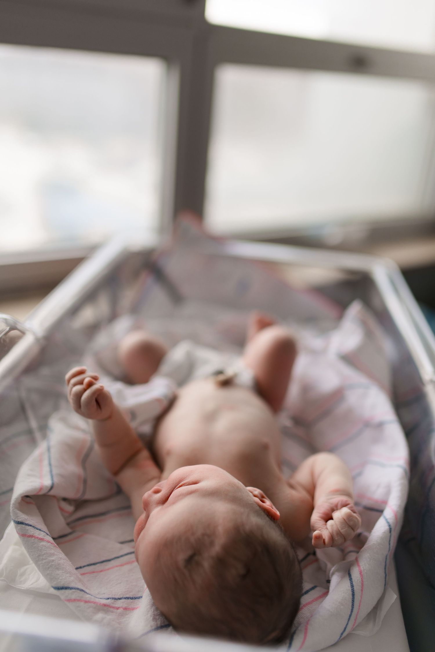 Newborn baby lying on striped blanket in hospital bassinet near window after birth.