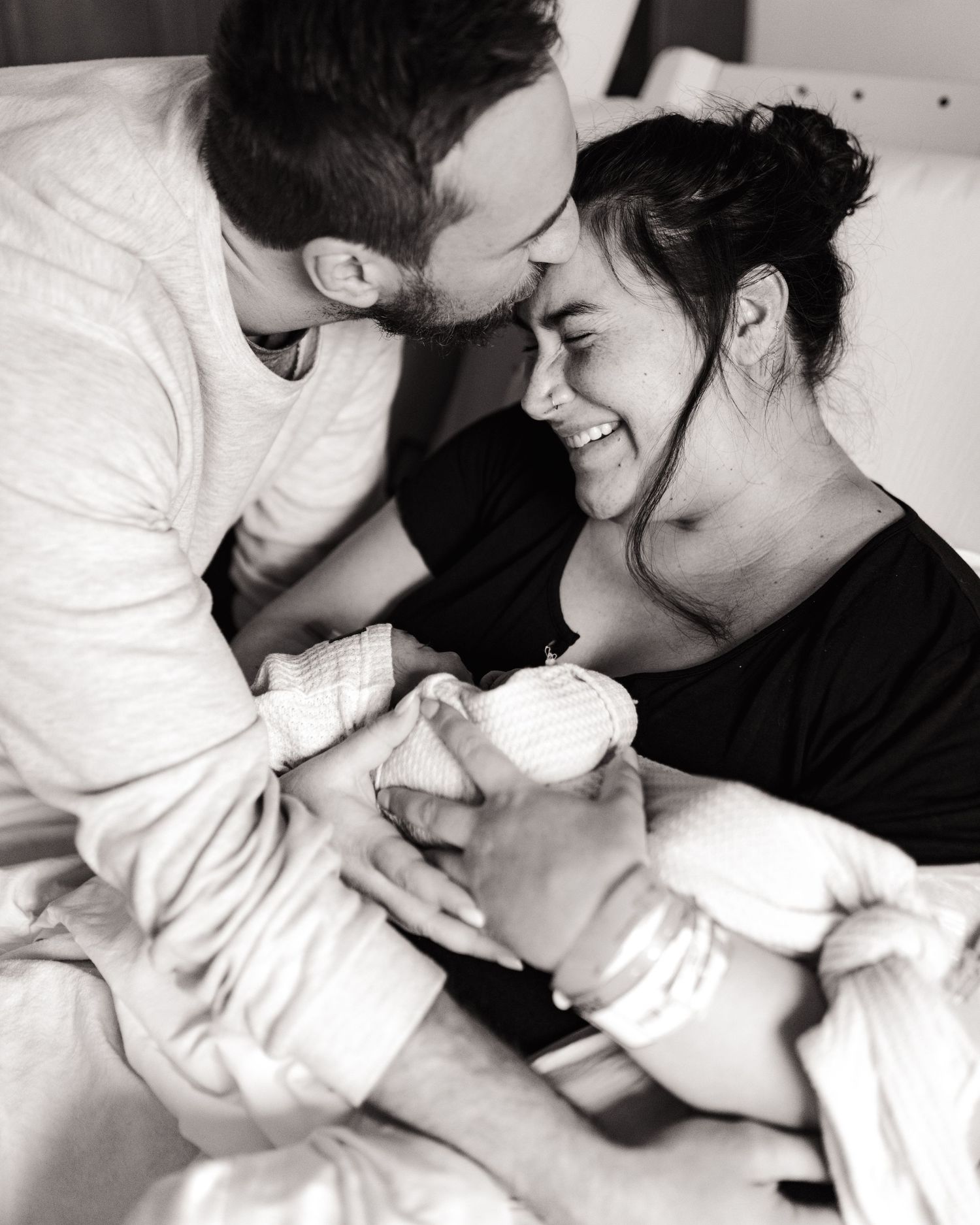 Black and white photo of new parents smiling and kissing their newborn baby in a hospital room.