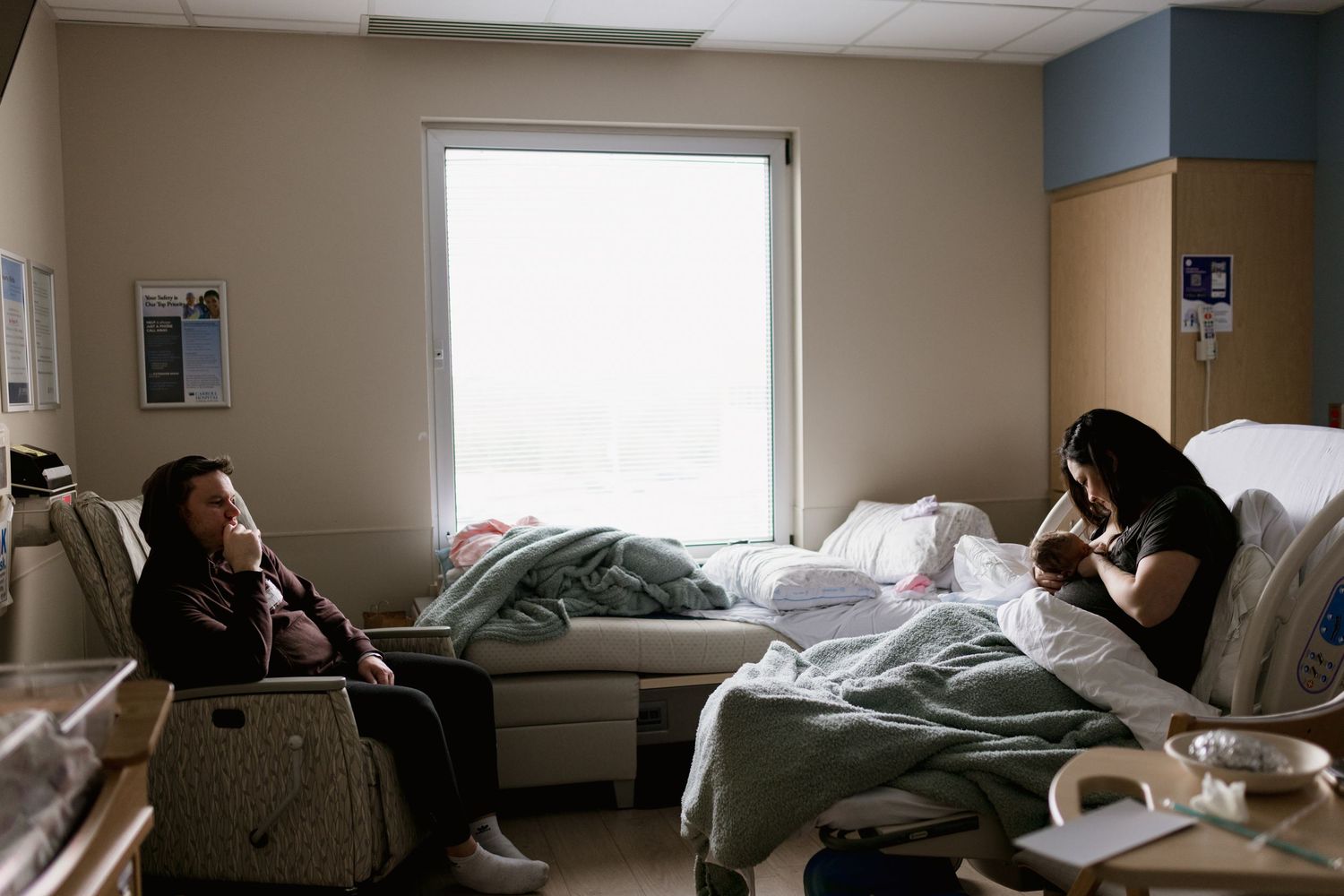 Two people in a hospital room, one resting in bed while the other sits nearby holding a newborn baby.