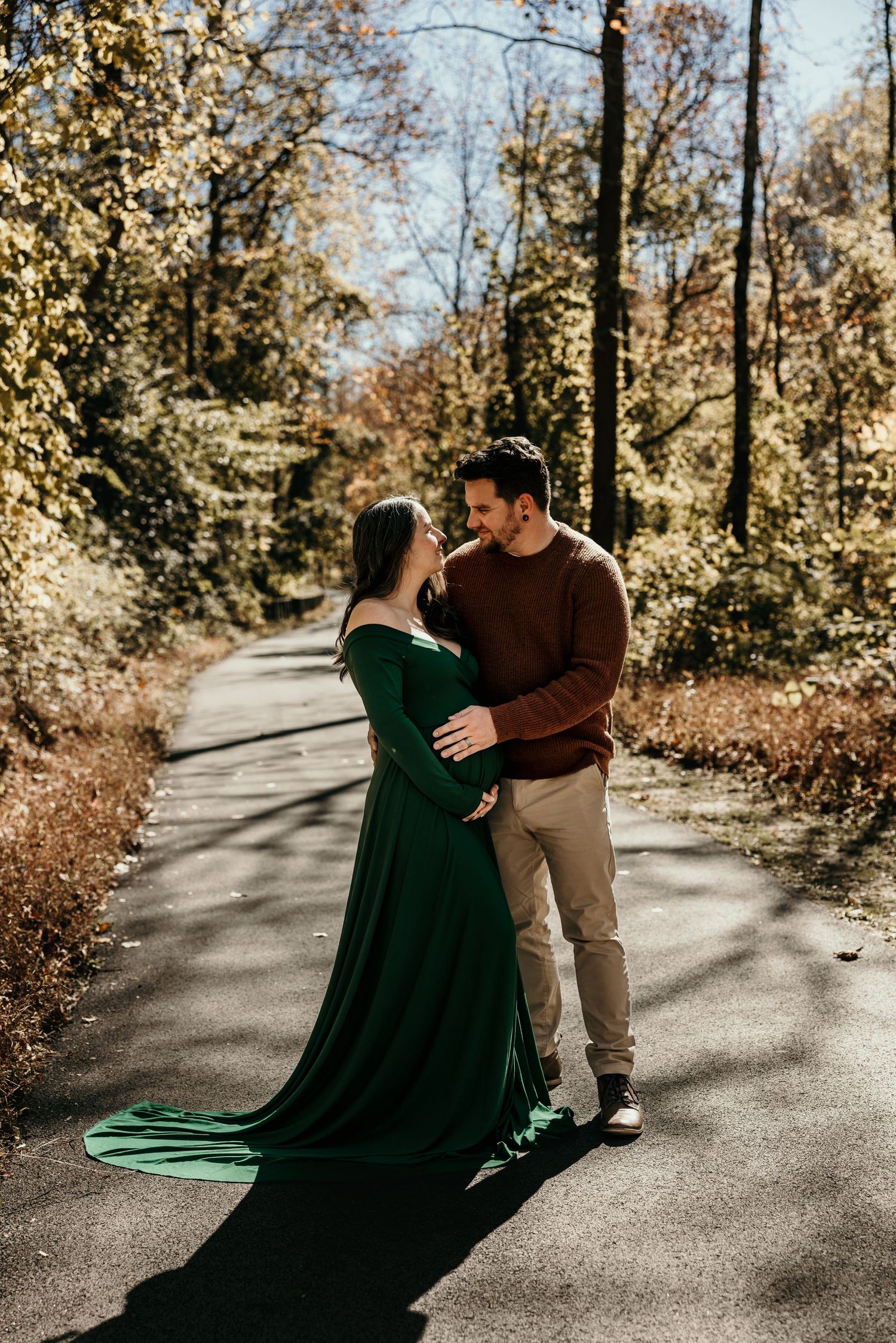 Couple embracing on sunlit autumn path, pregnant woman in elegant emerald green dress with long train.