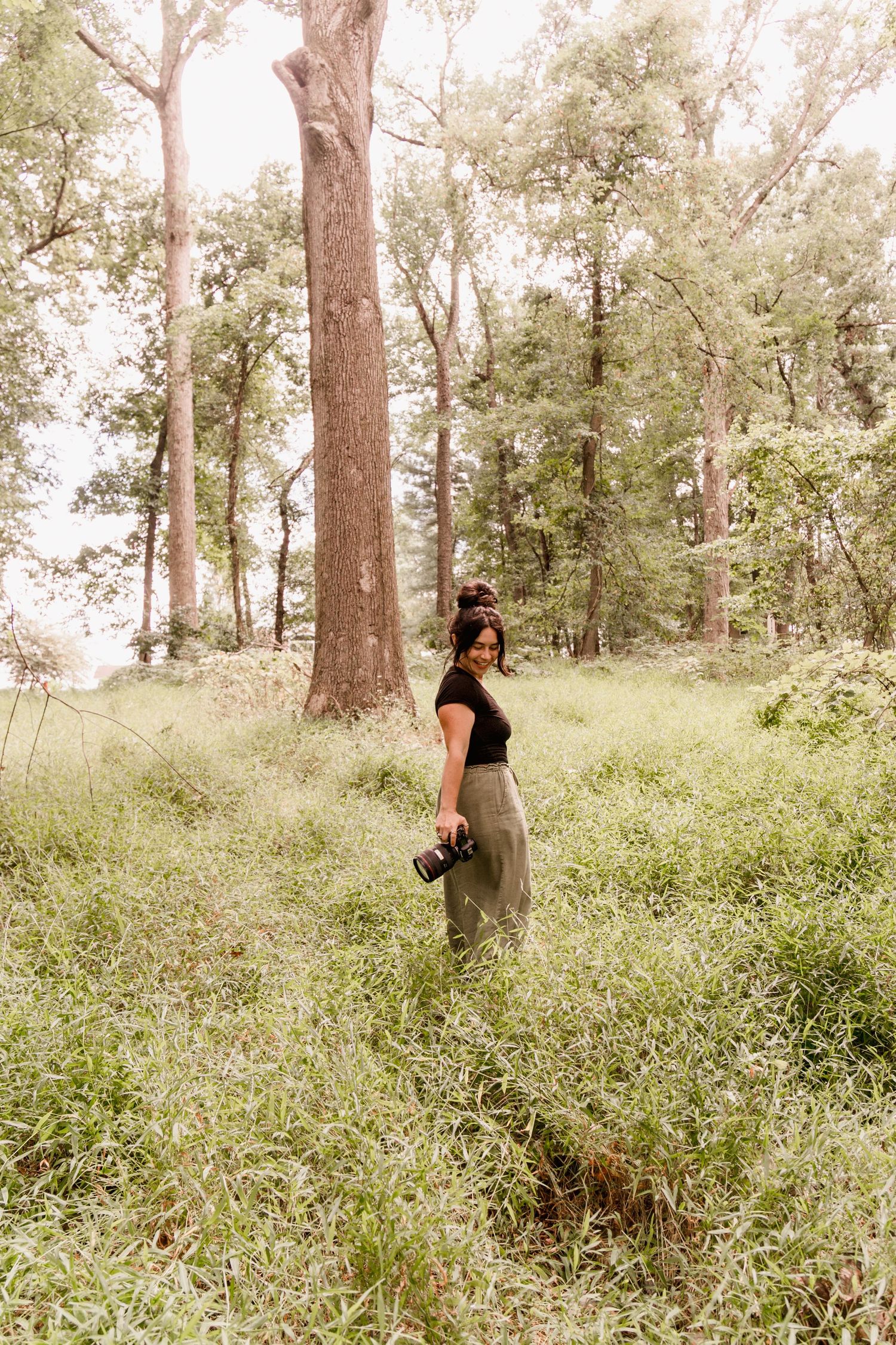 Woman in black top and olive pants holding a camera while standing in a lush green forest clearing.