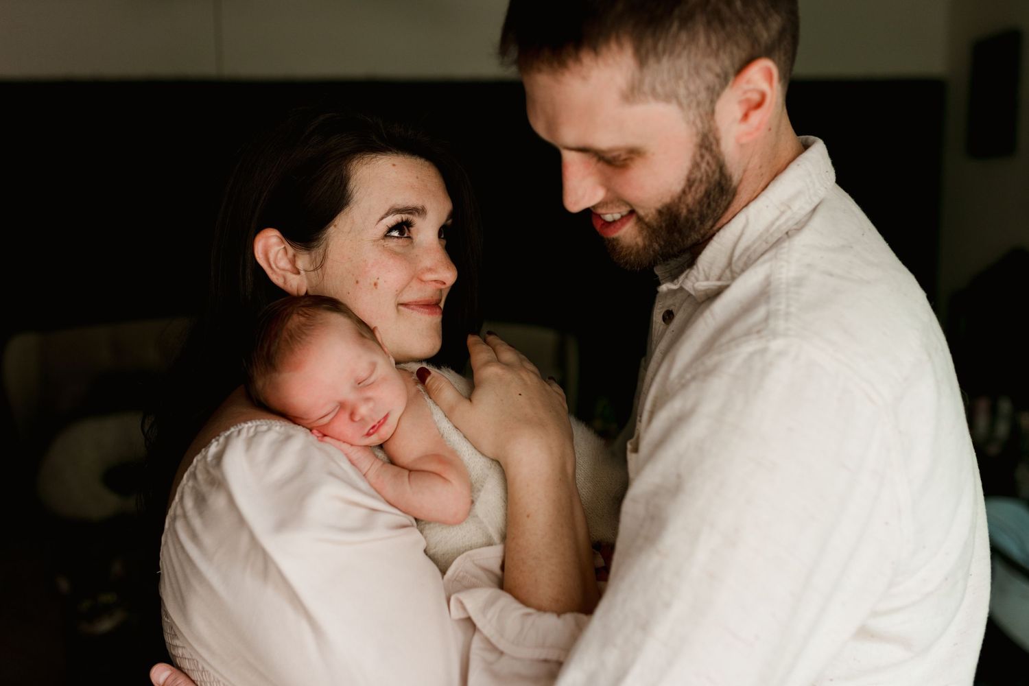 New parents share a tender moment holding their sleeping newborn baby in a softly lit room.