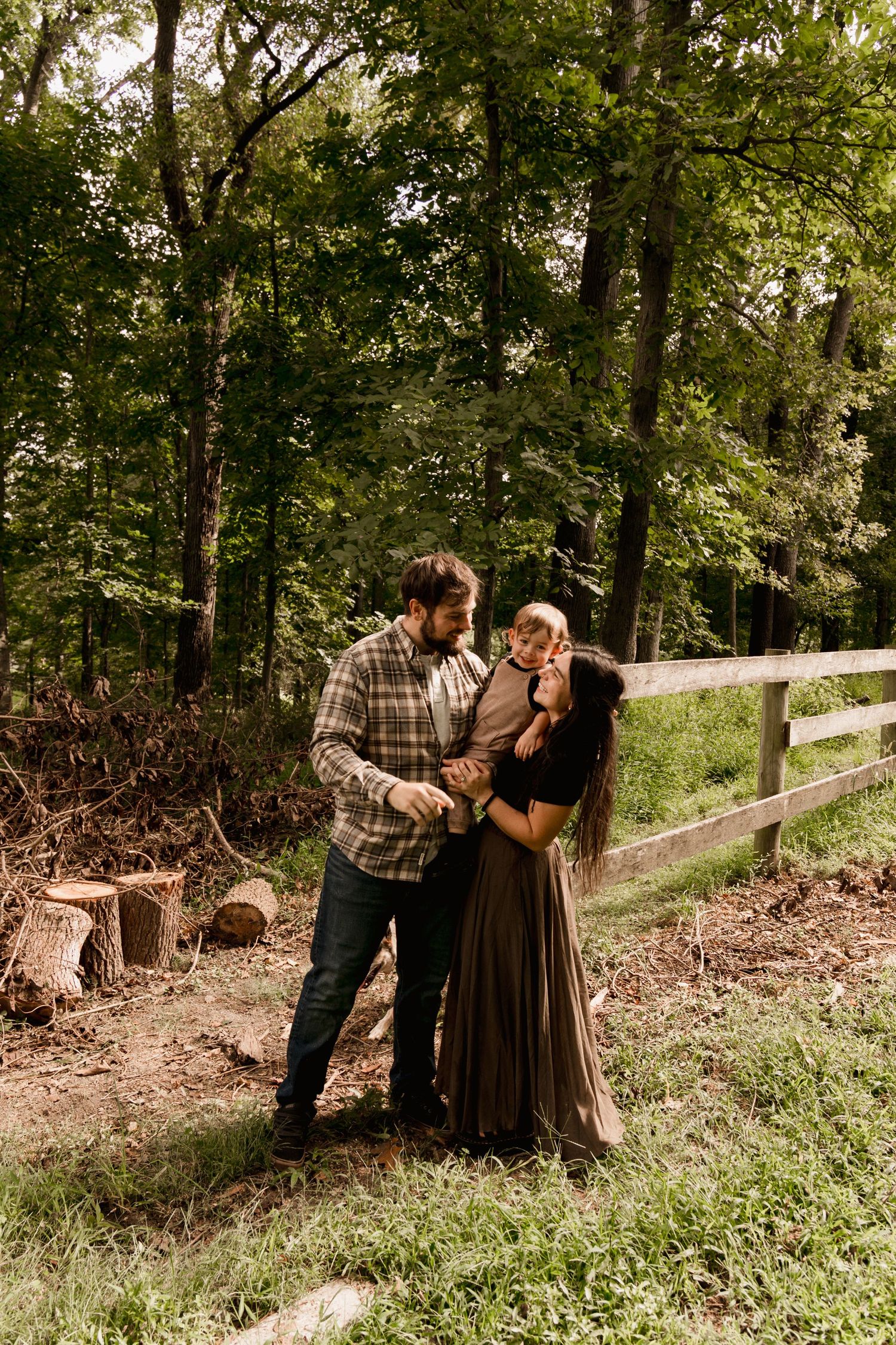 A couple stands together in a wooded area, the woman holding a baby while the man looks on tenderly.
