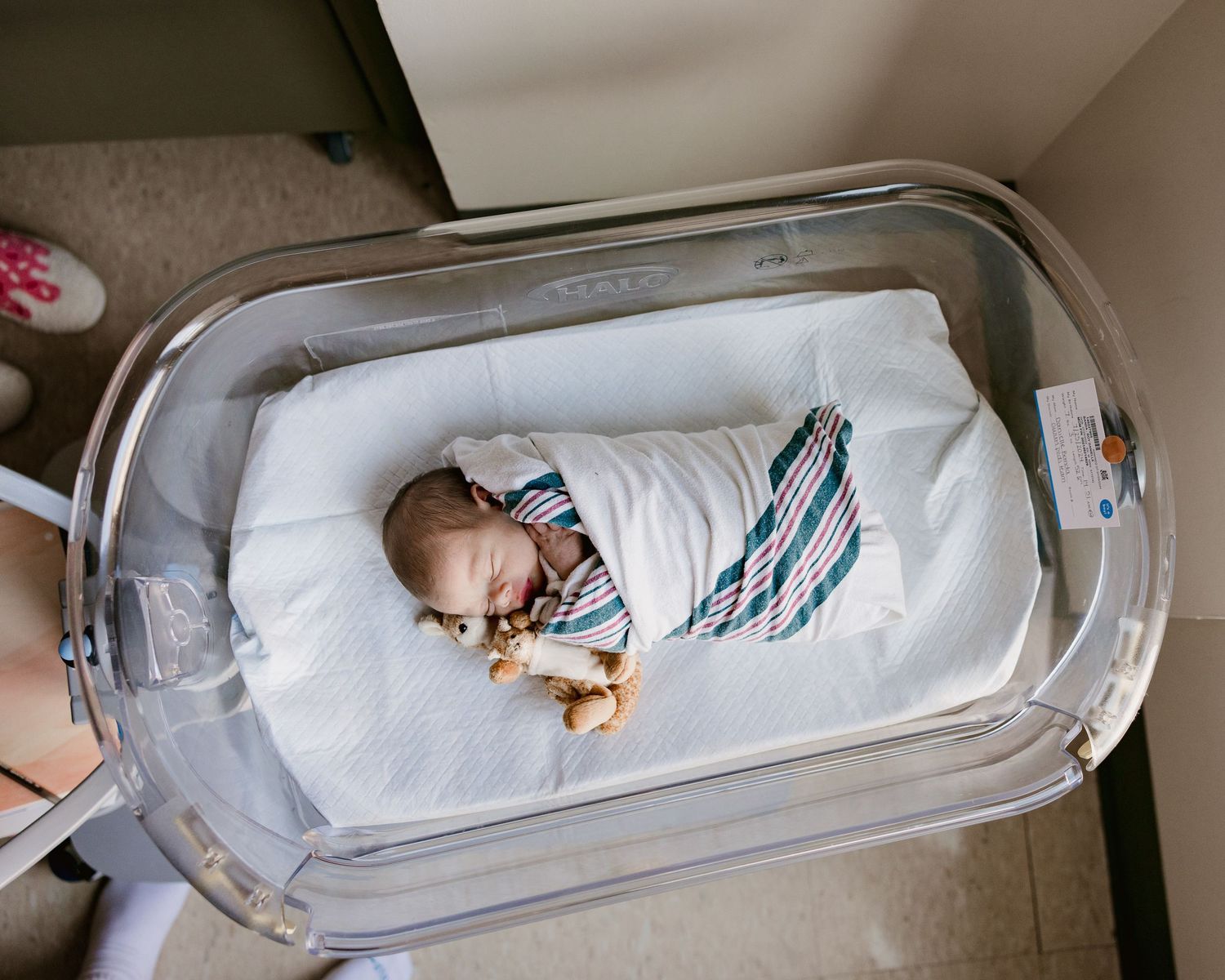 Newborn baby swaddled in striped blanket sleeping peacefully in a clear hospital bassinet.