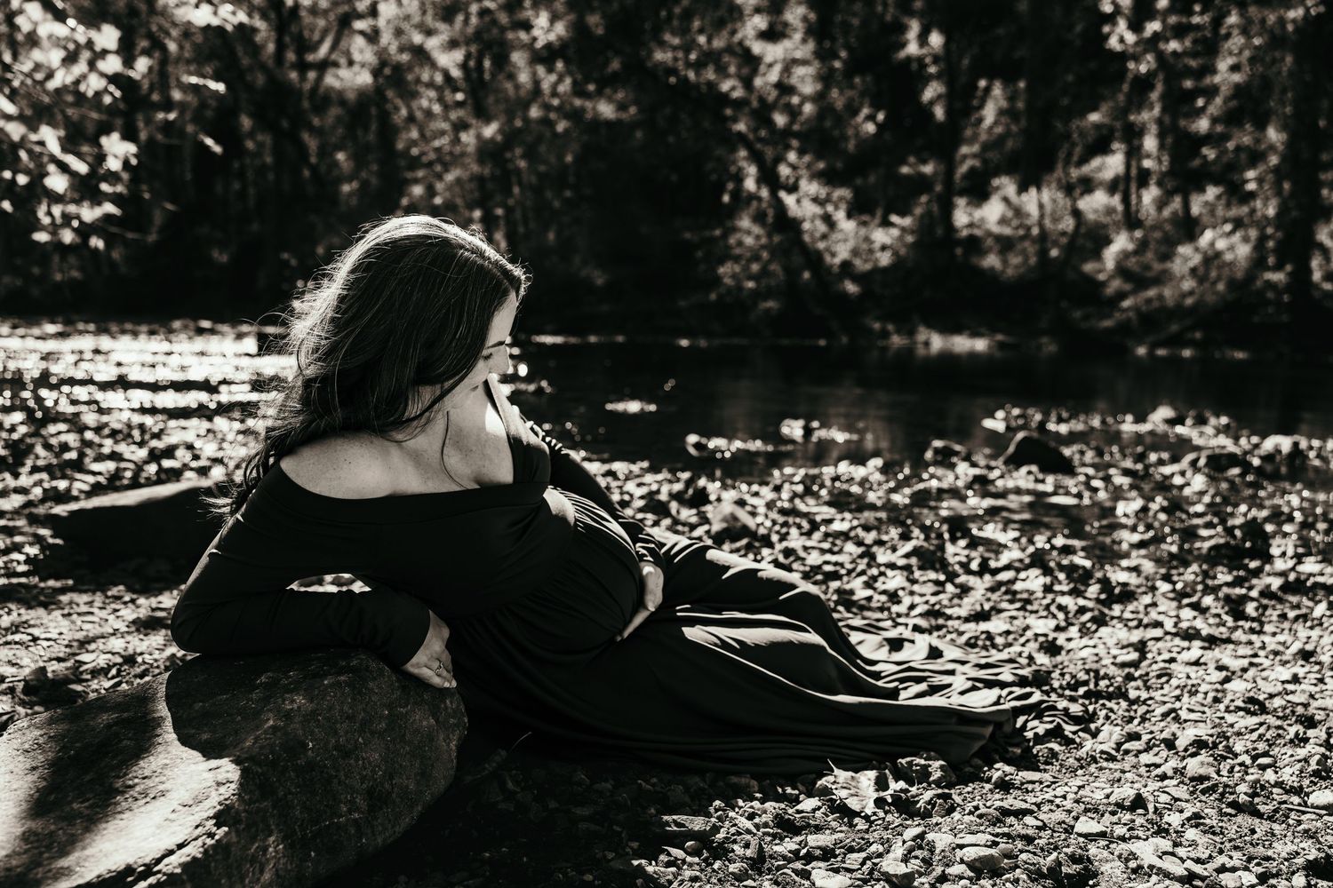Pregnant woman in dark off-shoulder gown reclining on rocks near creek in black and white maternity photo.