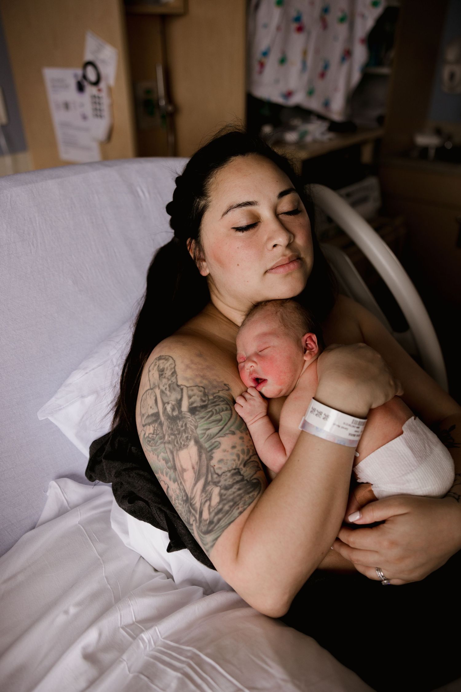 New mother with tattoos holds sleeping newborn skin-to-skin in hospital bed after birth.