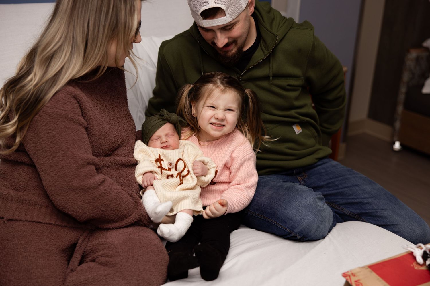 A family of four poses in a hospital room, with parents and toddler girl smiling while holding their newborn baby.