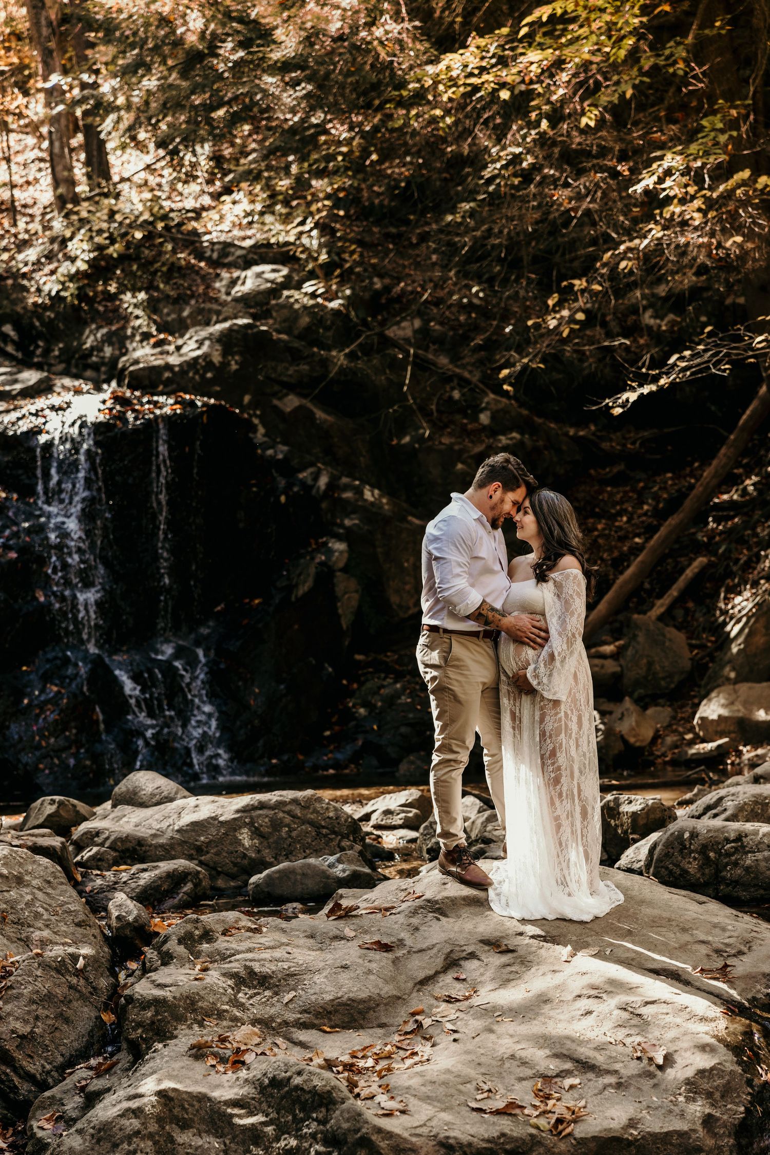 Couple embracing romantically on rocks before a waterfall, pregnant woman in sheer white lace maternity gown.