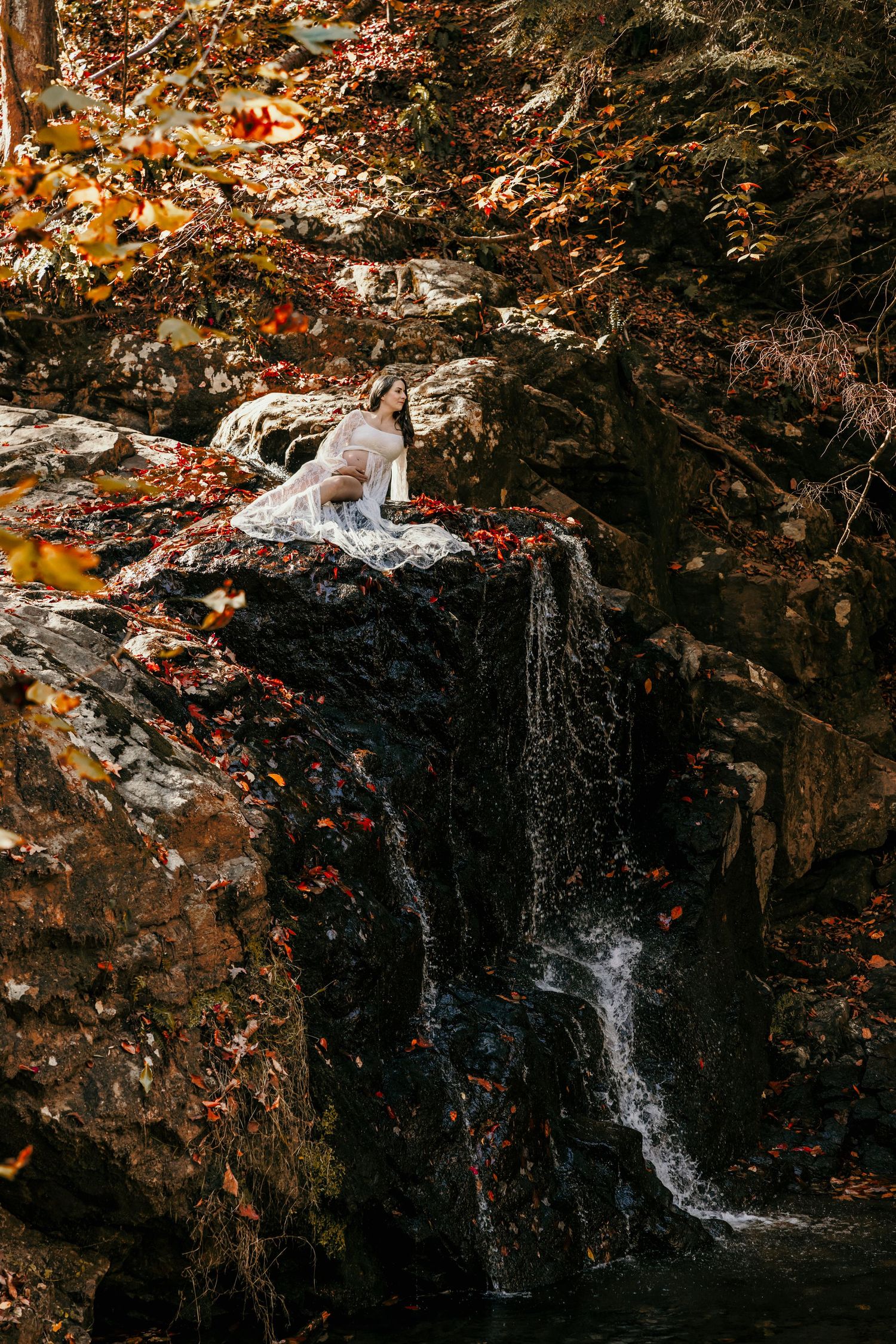 Woman in white dress sitting on rocky ledge beside cascading waterfall with autumn foliage backdrop.
