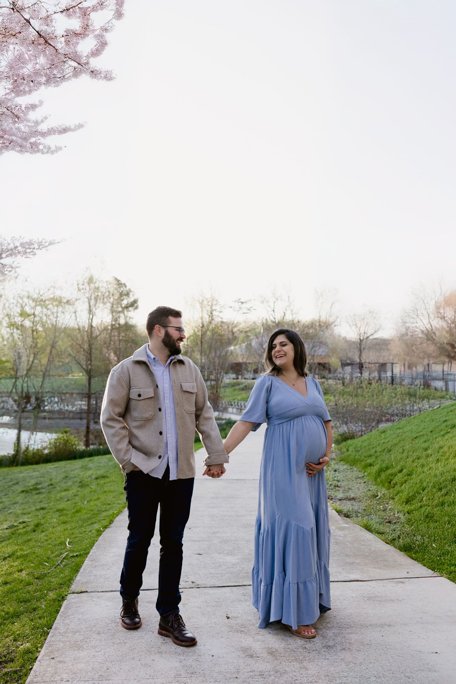 Expecting couple holds hands walking on a path surrounded by blooming cherry blossom trees in spring.
Storyteller Photographer