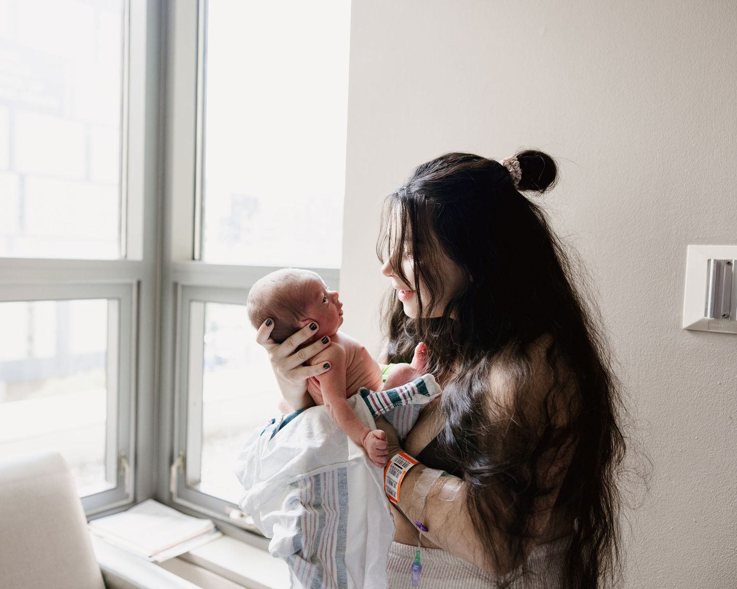 A mother with long dark hair holds her newborn baby up near a bright hospital window.