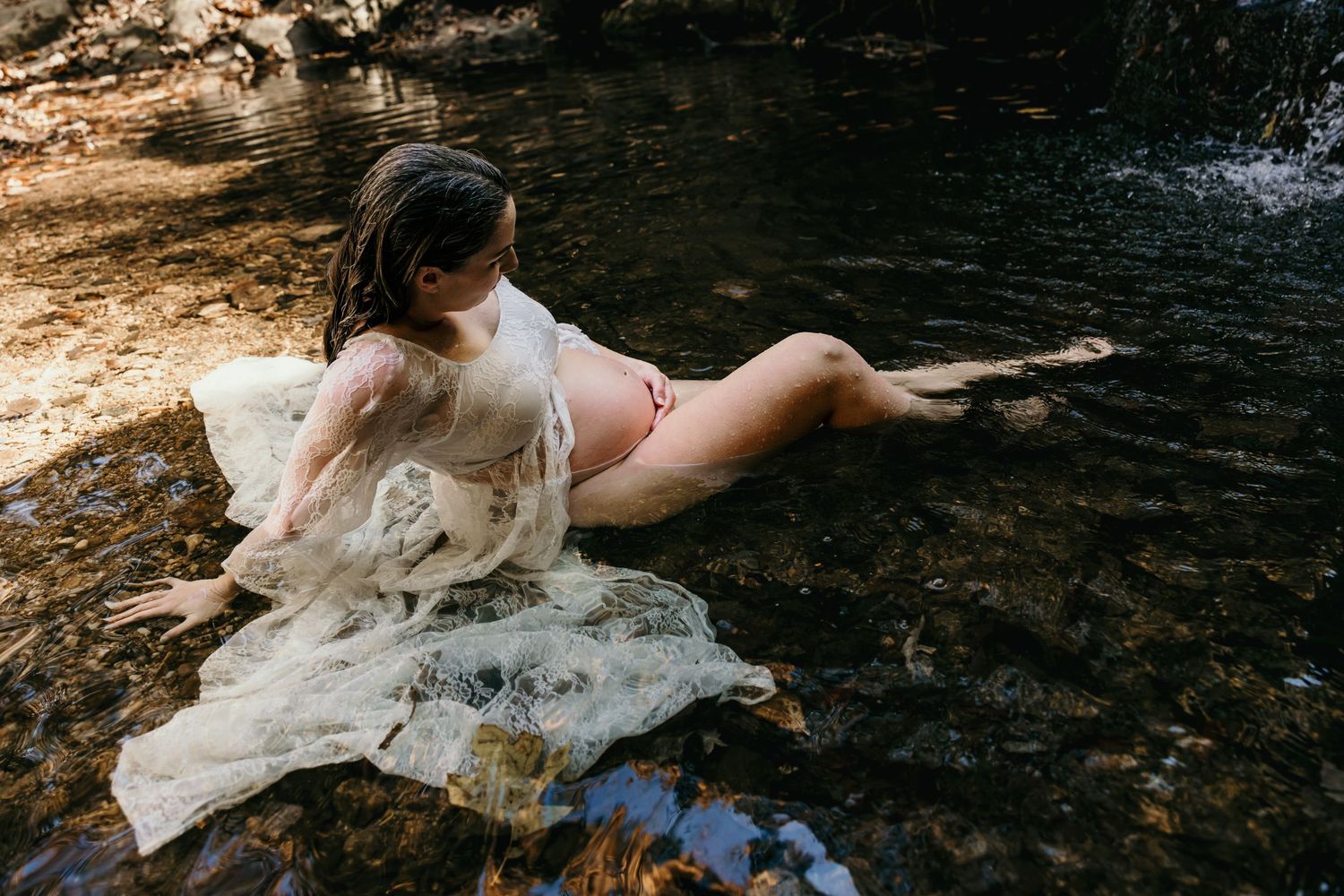 Pregnant woman in sheer white lace gown reclining gracefully in shallow creek surrounded by rocks.