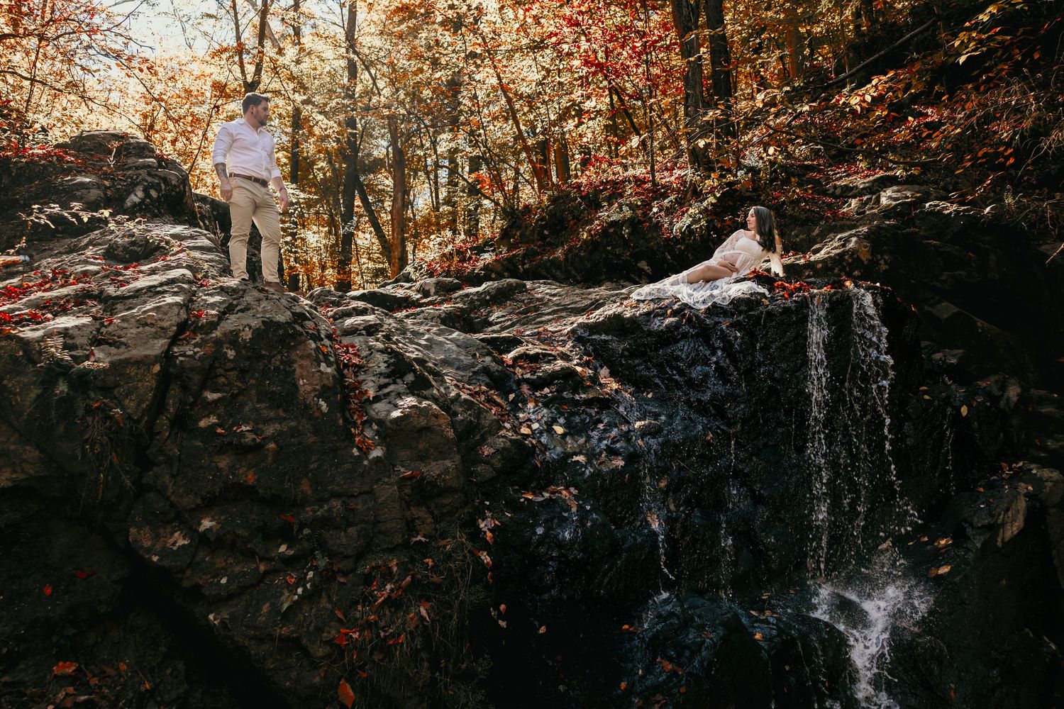 Couple standing on rocky ledge beside a waterfall in autumn forest, woman in flowing white dress.
