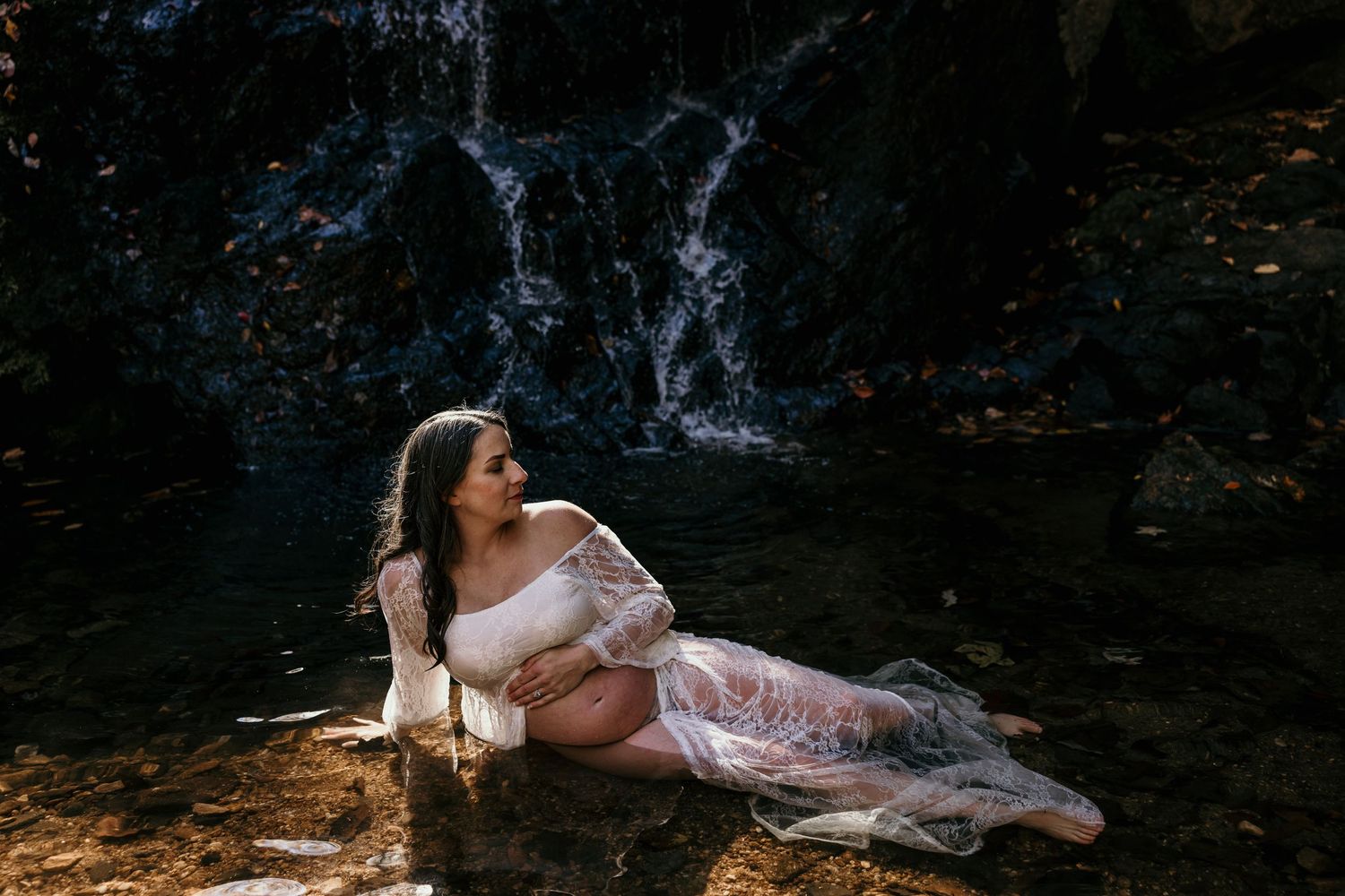 Pregnant woman in sheer white lace gown reclining in shallow water before a rocky waterfall.