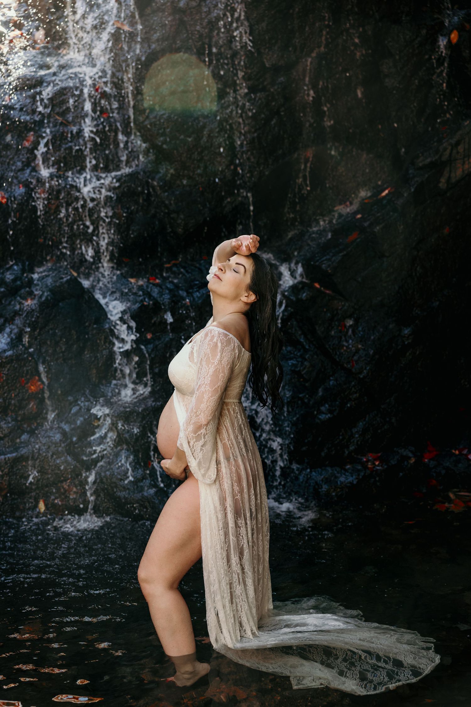 Pregnant woman in sheer white lace gown standing in water before a rocky waterfall in autumn.
