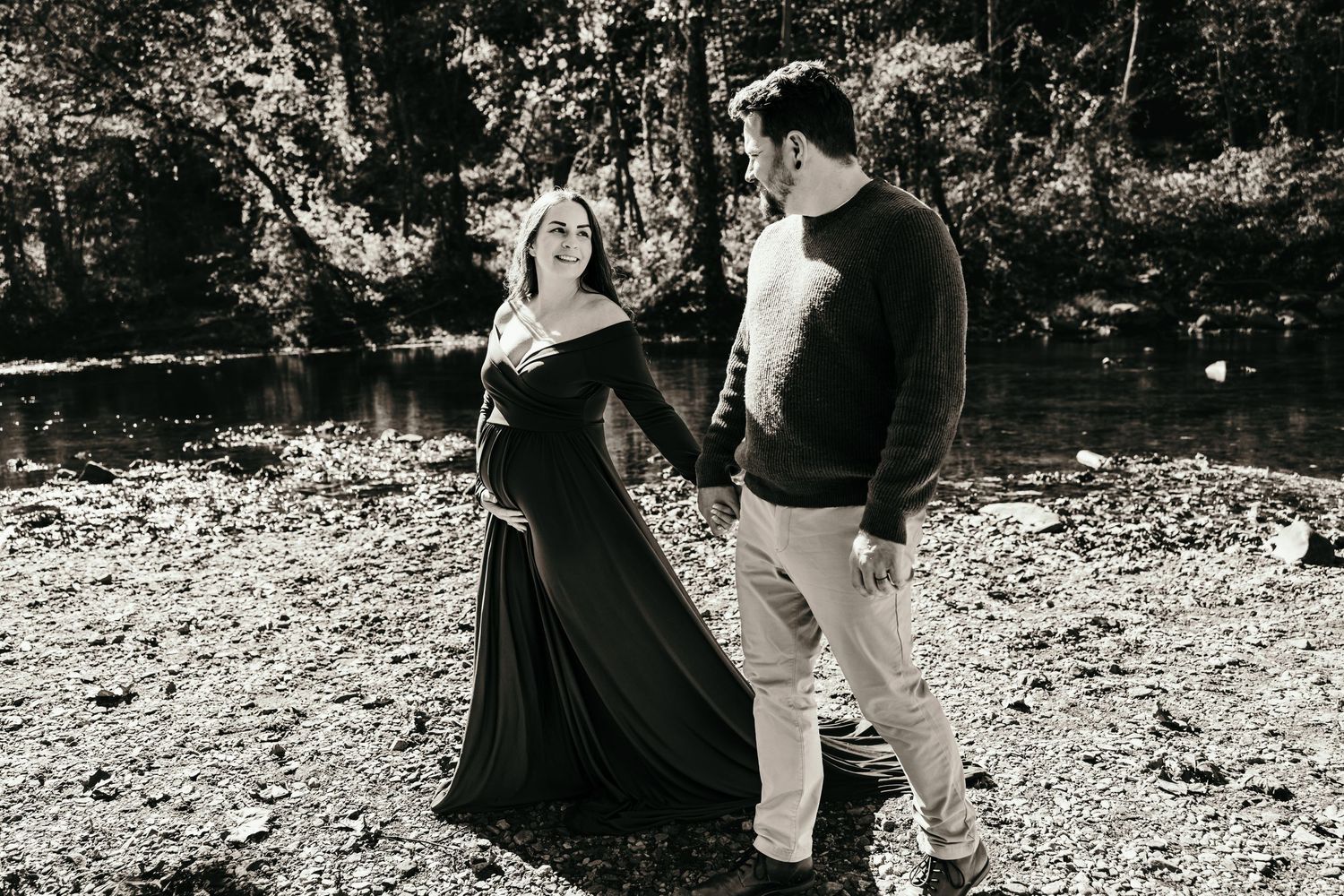Couple holding hands near forest creek, pregnant woman in dark flowing gown, black and white maternity portrait.