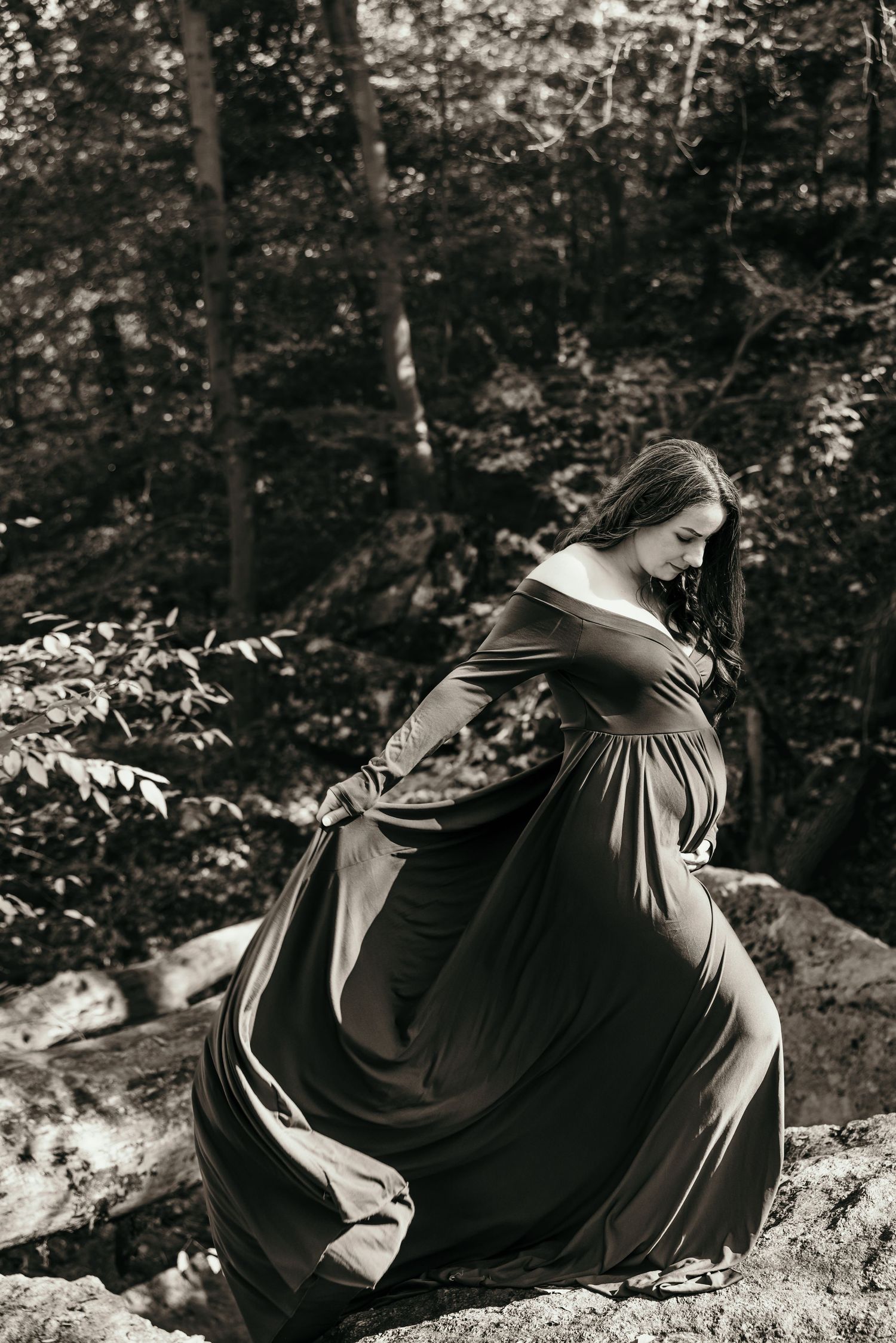 Pregnant woman in flowing dark off-shoulder gown standing on rocks in forest, black and white maternity photo.