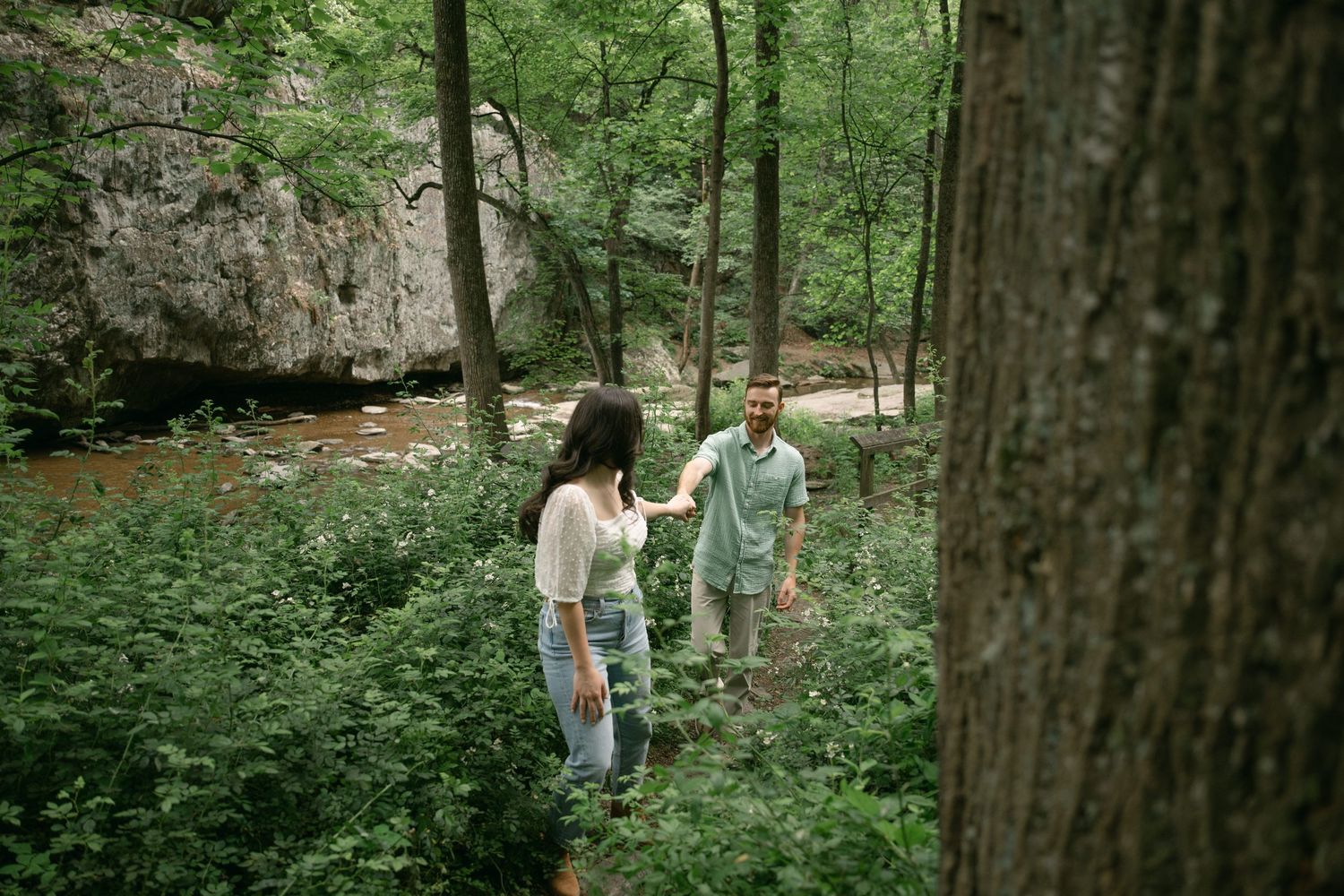 Couple walking through a lush green forest path surrounded by tall trees.