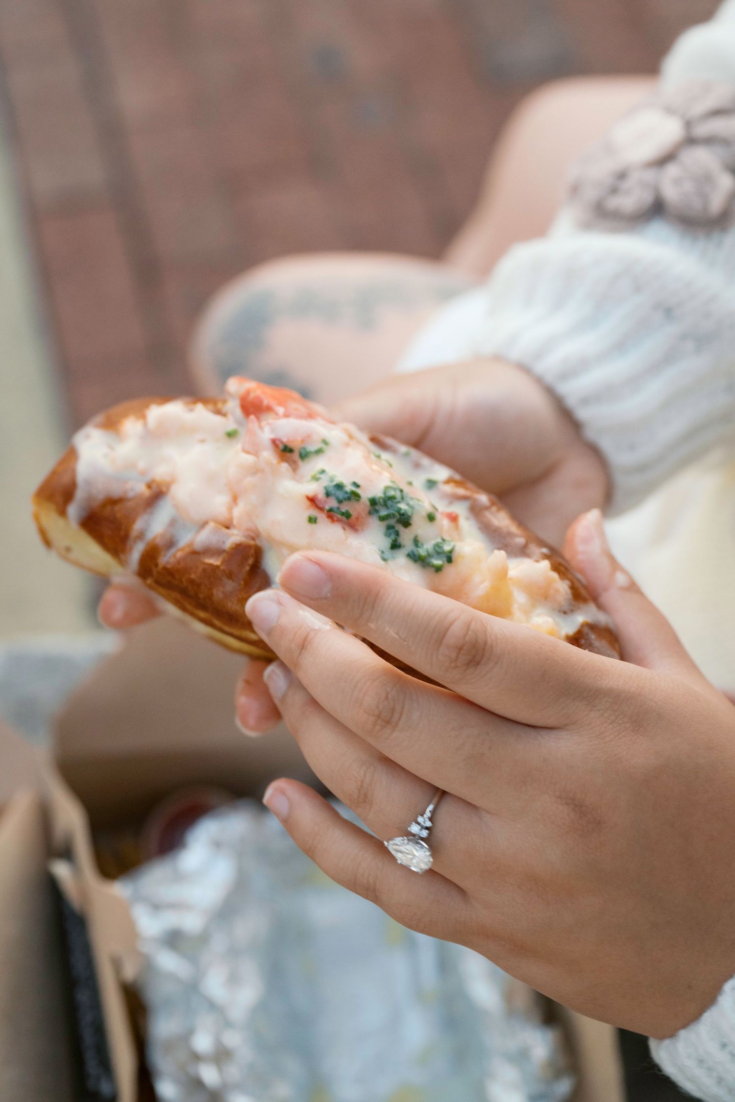 Hands holding a buttery lobster roll with garnish and wearing an engagement ring on brick background.