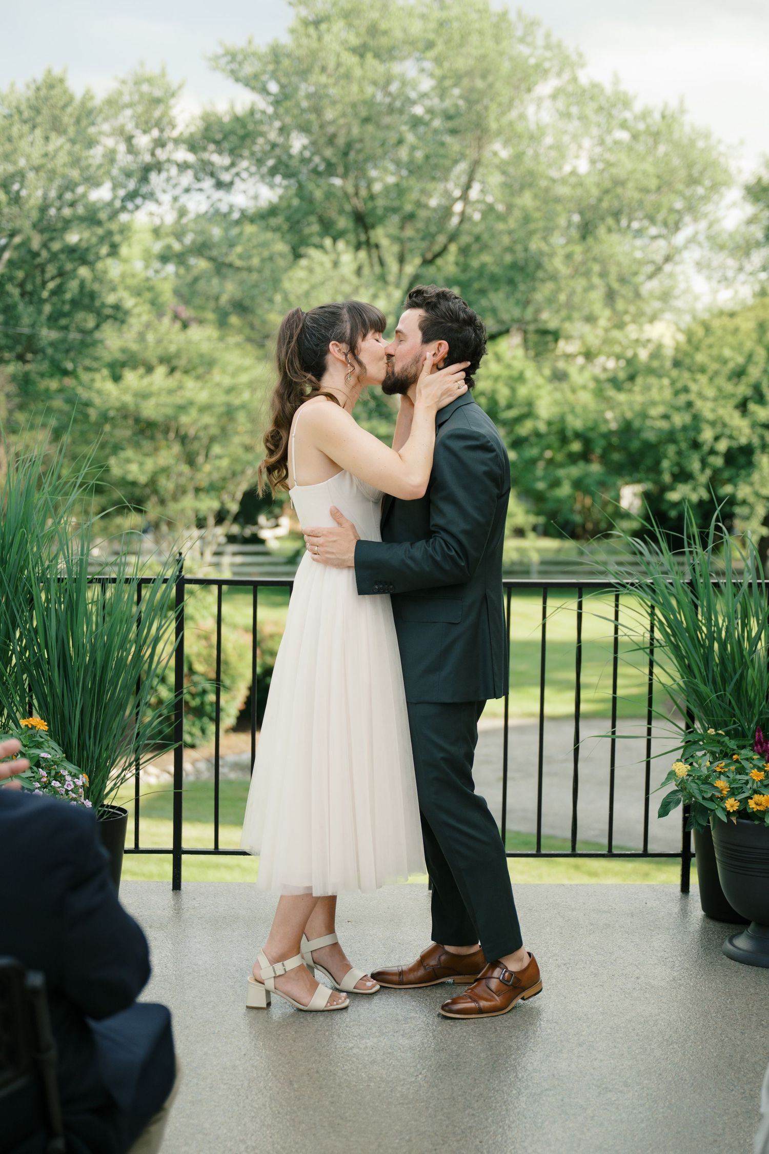 Wedding ceremony kiss on a patio with green trees and decorative plants in the background.
