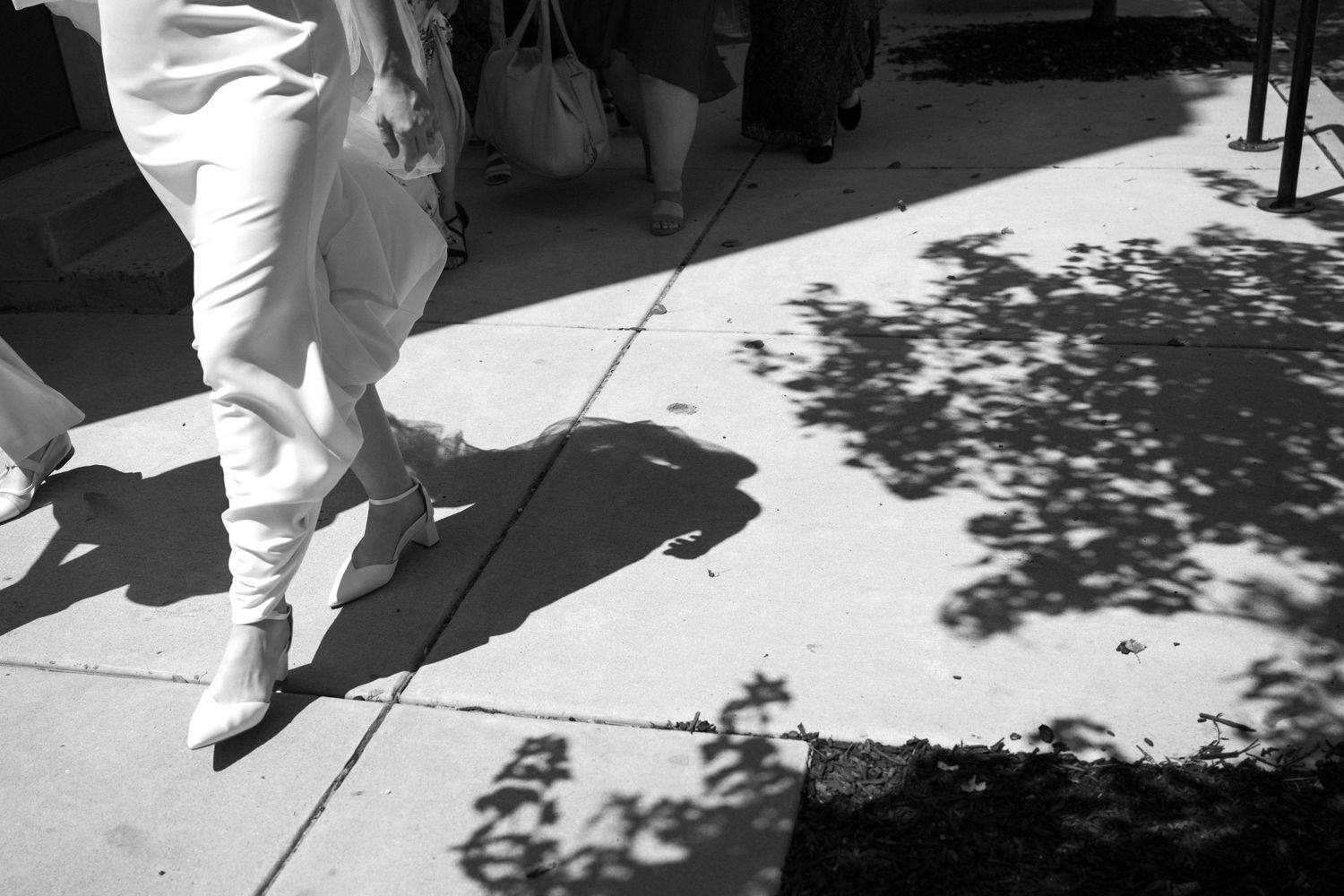 Black and white street photography showing tree shadows cast on a sidewalk with someone wearing white clothing walking.