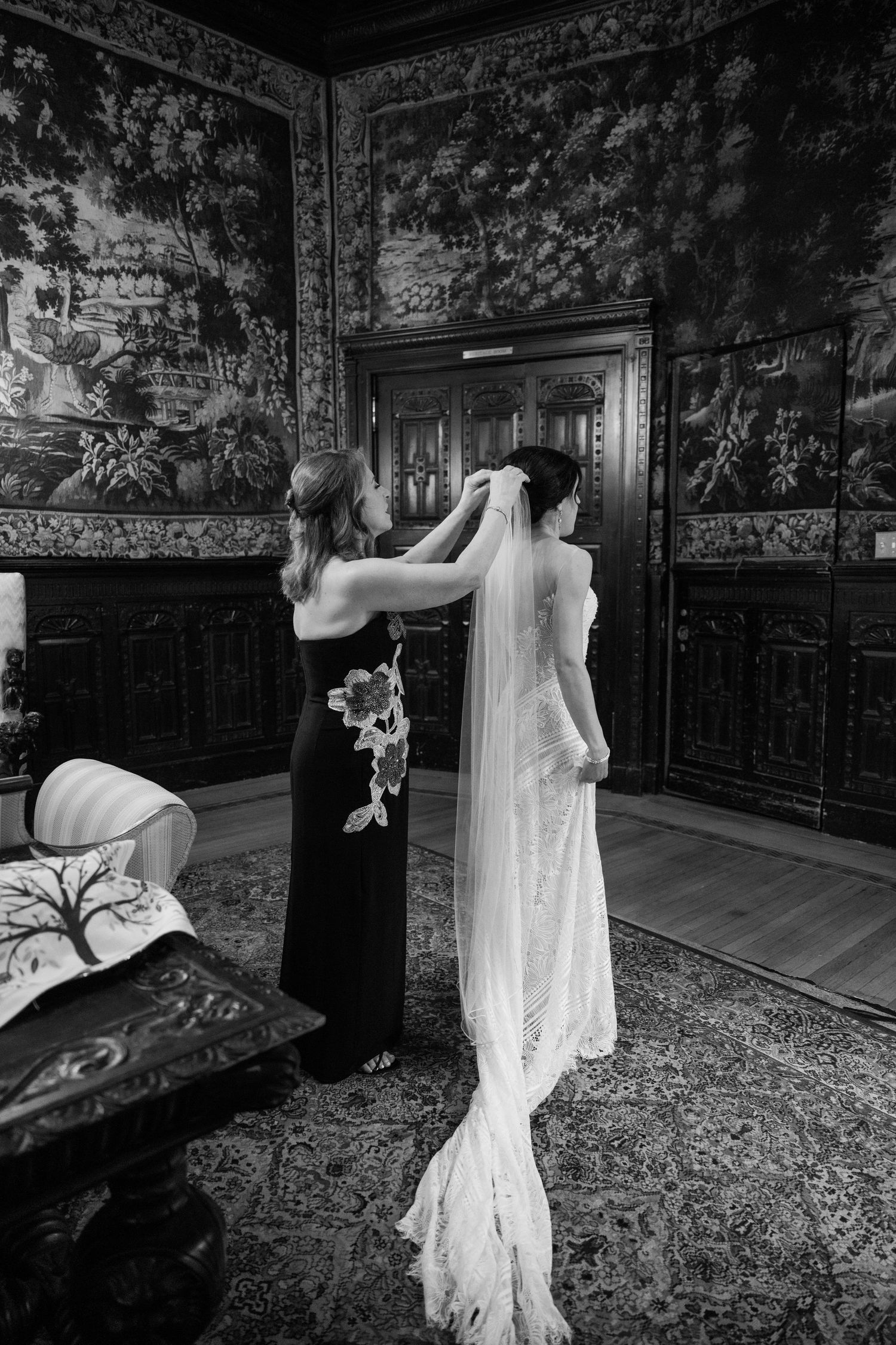 Black and white photo of a wedding dress being adjusted in an ornate room with tapestry walls and antique decor.