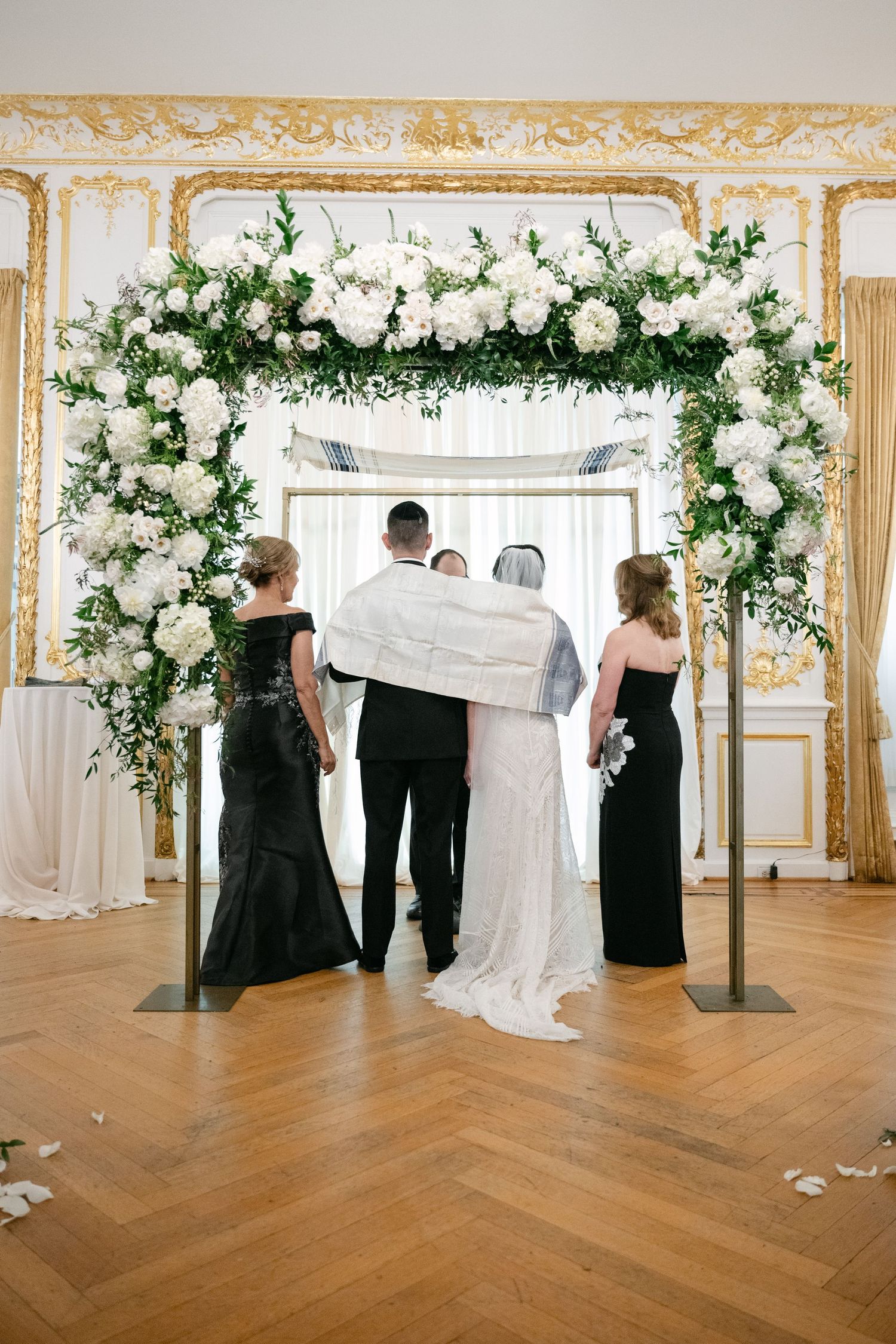 Wedding ceremony under floral arch with white roses and greenery in elegant gold-trimmed room.