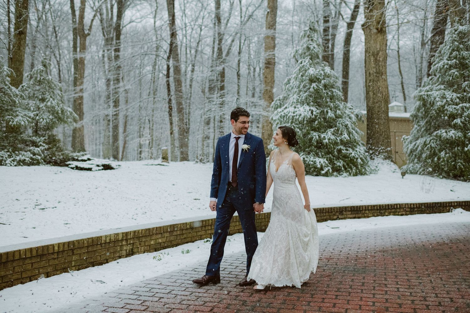A couple walks along a snowy brick path surrounded by winter trees.